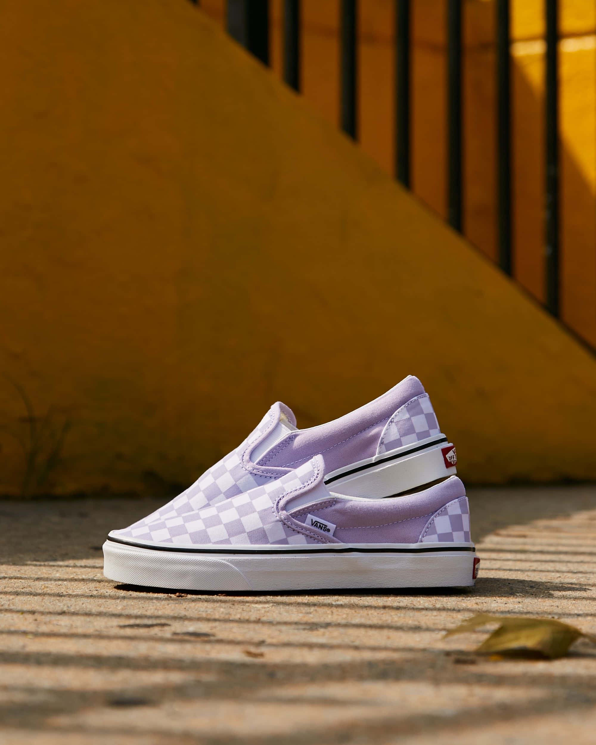 Vans 桌布 壁紙 — 兩雙薰衣草色格紋Vans slip-on鞋放在陽光照射的混凝土上，背景是溫暖的黃色牆壁和黑色欄杆。