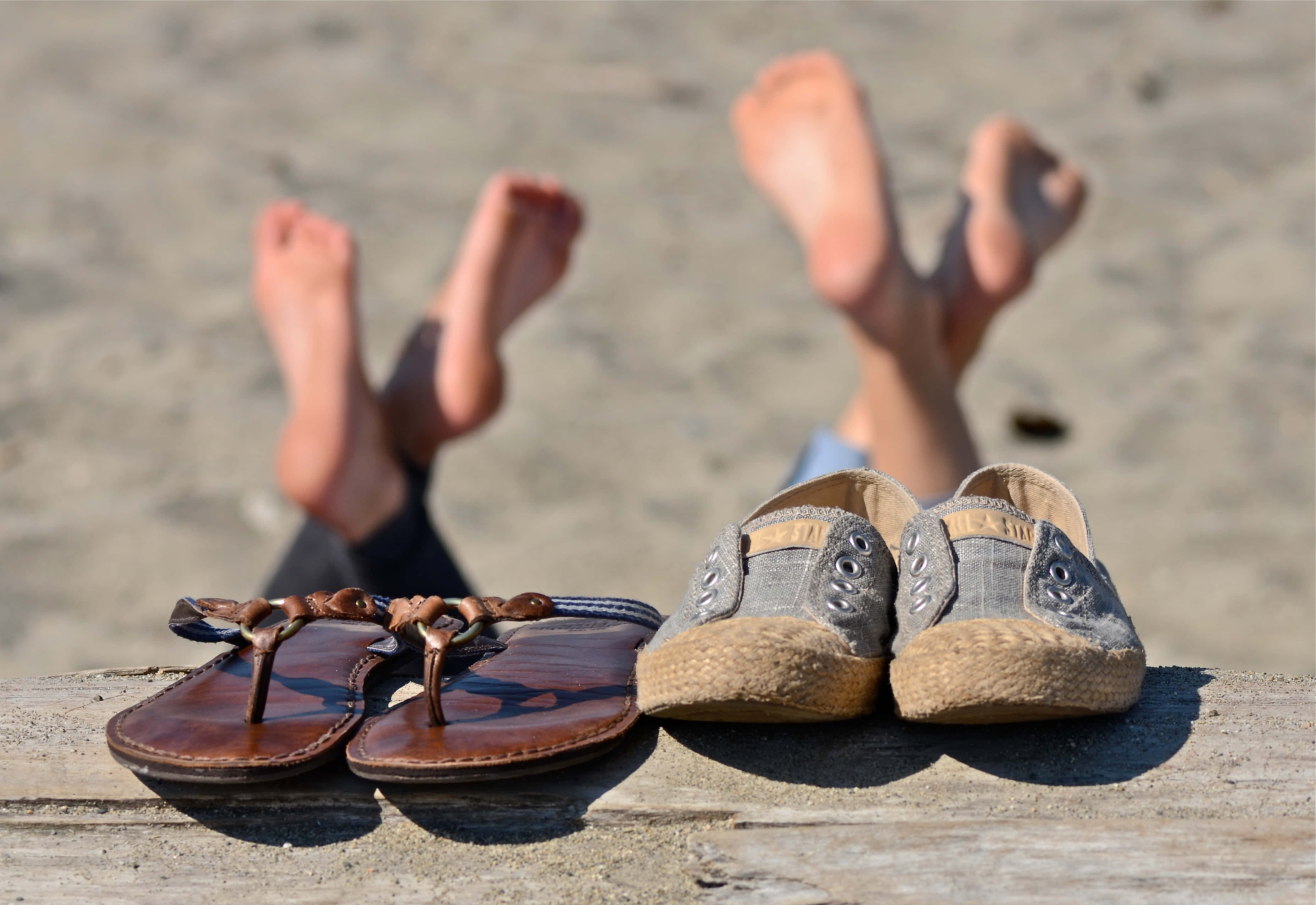 Beach Sandals 桌布 壁紙 — 兩雙鞋放在一塊木板上，赤腳在陽光明媚的海灘上踢起沙子。