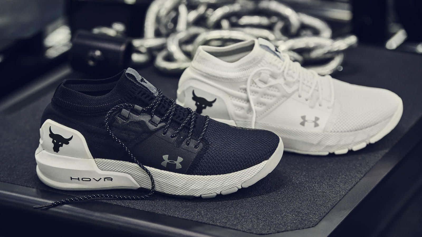 Project Rock 桌布 壁紙 — 兩雙黑色和白色的Under Armour HOVR運動鞋，帶有公牛標誌，放在健身墊上，旁邊有盤繞的繩索和啞鈴。