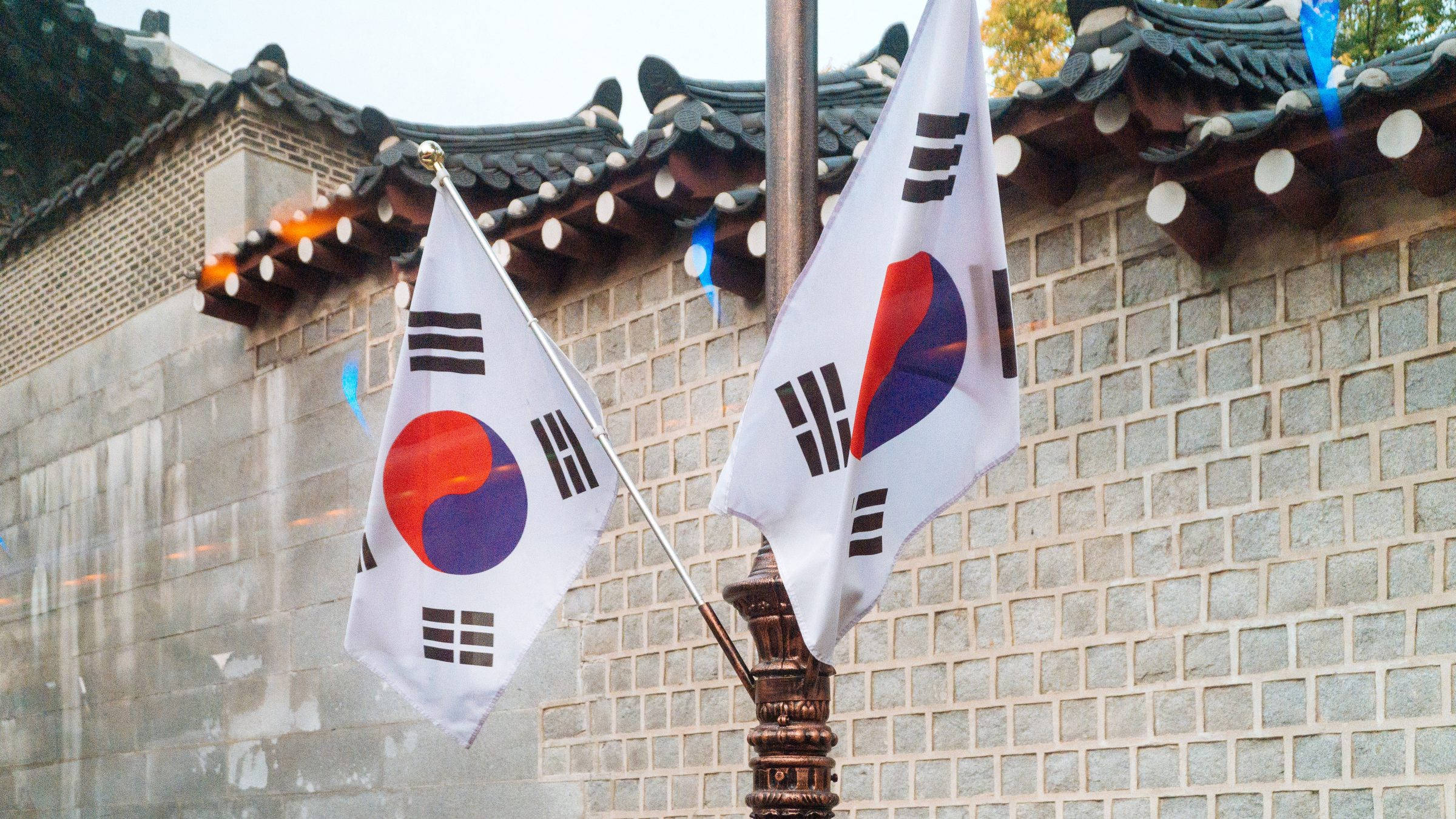 Flag 桌布 壁紙 — 兩面韓國國旗在傳統瓦頂和石牆前飄揚，柔和日光下。