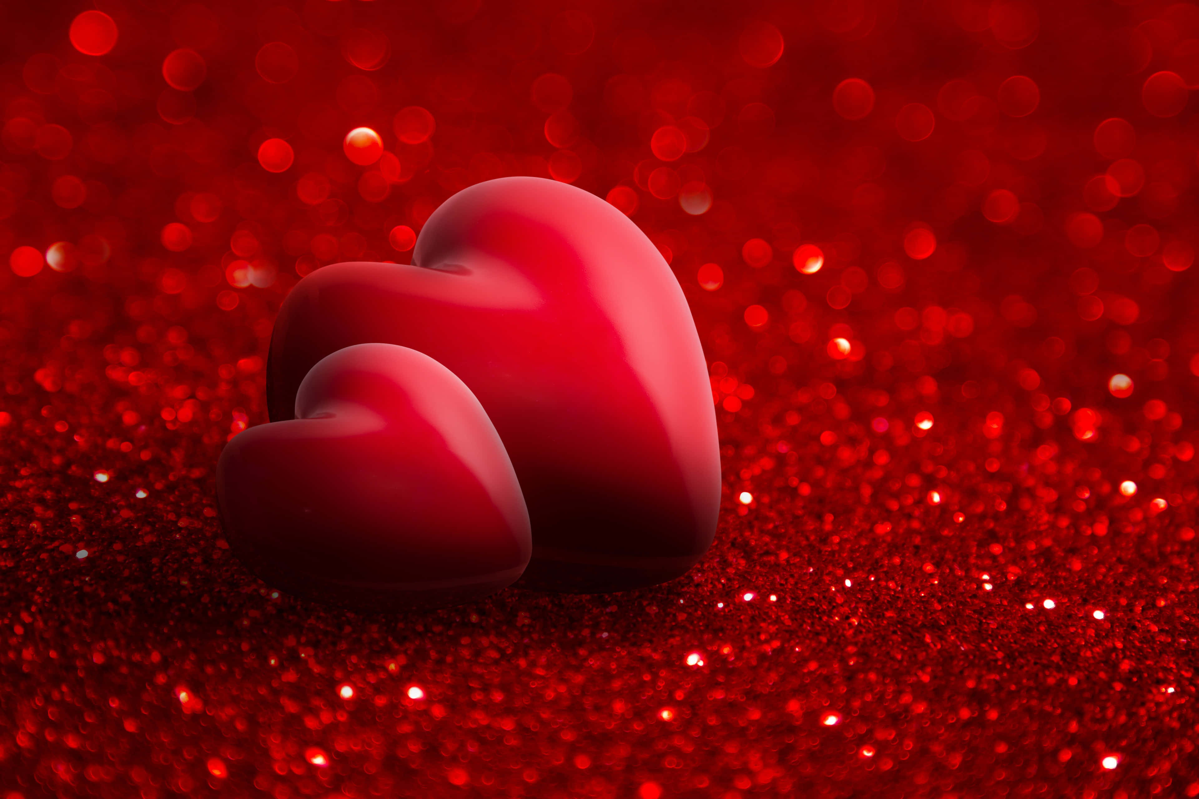 3d Heart 桌布 壁紙 — 兩顆光滑的紅色心形放在閃閃發光的紅色亮粉表面上，背景有柔和散景燈光。