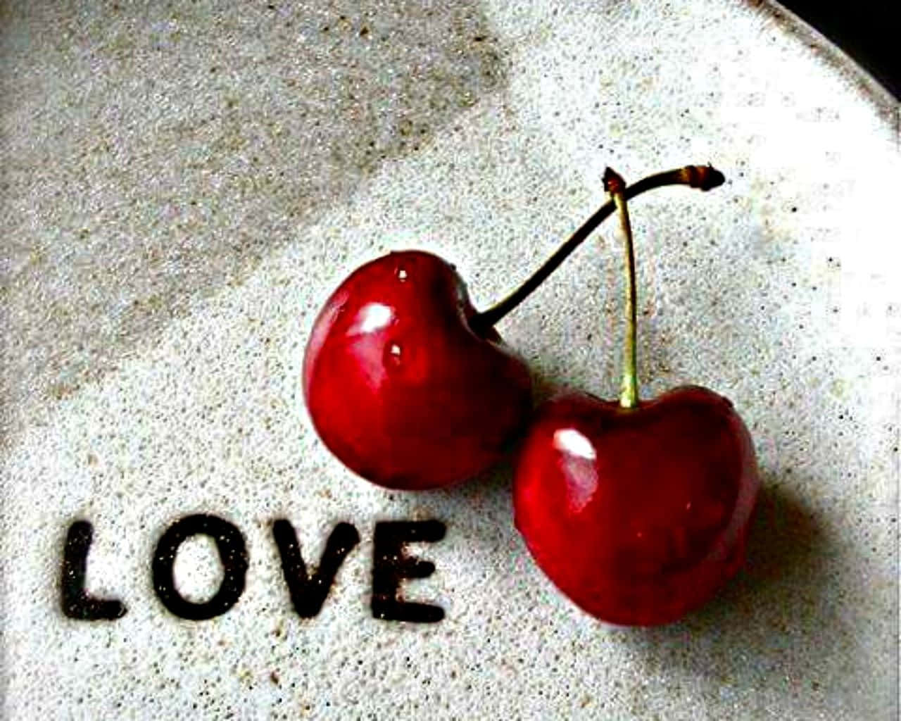 Cherry Aesthetic 桌布 壁紙 — 兩顆光澤的紅色櫻桃放在一個斑點白色表面上，旁邊是用深色粉末蝕刻的LOVE字樣。