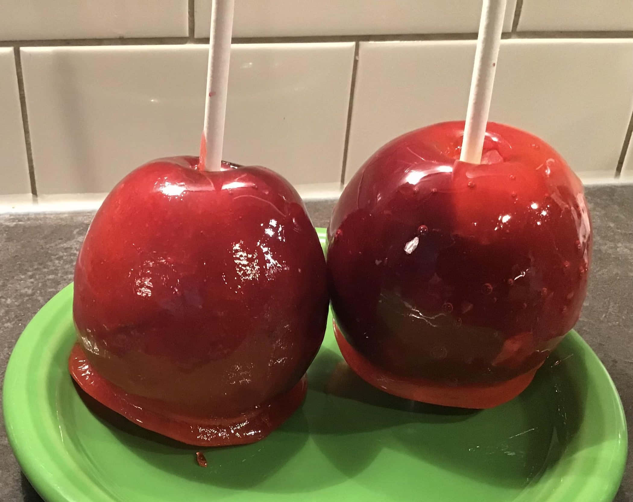 Candy Apples 桌布 壁紙 — 兩顆光澤的紅色糖蘋果插在棒上，放在一個亮綠色盤子上，背景是瓷磚廚房。