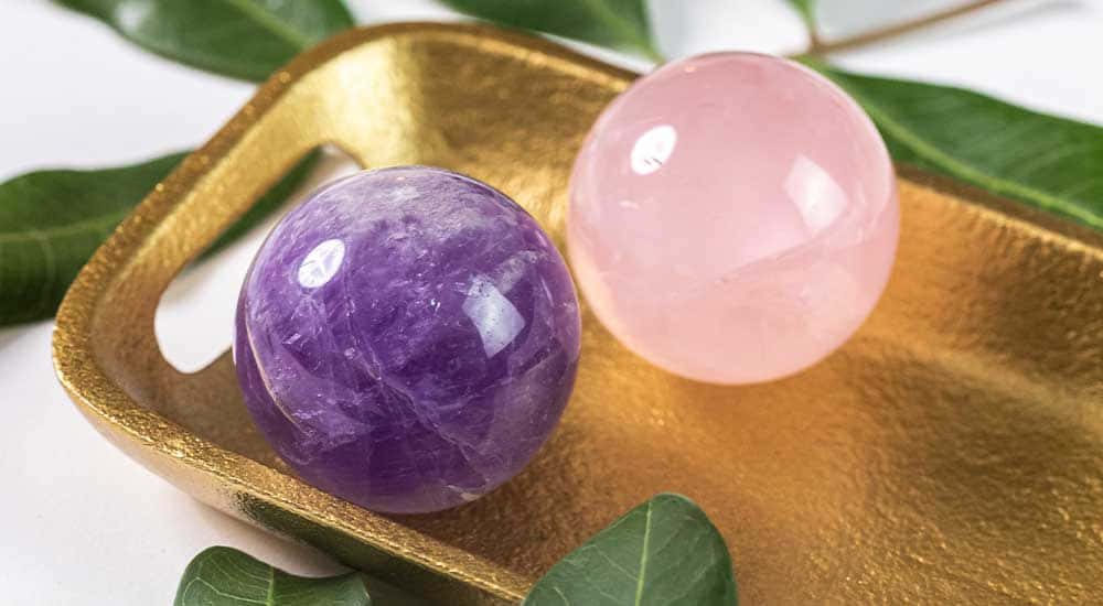 水晶 桌布 壁紙 — 兩顆拋光寶石球體，紫色紫水晶和粉紅玫瑰石英，放在一個被綠葉環繞的金色托盤上。