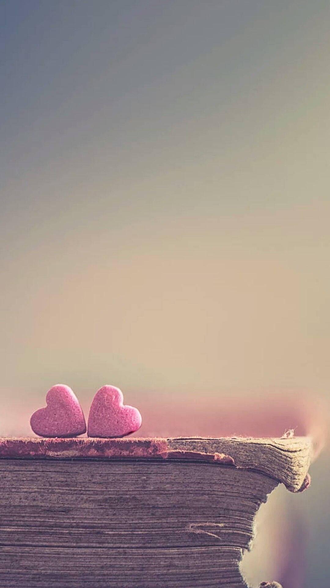 Love Phone 桌布 壁紙 — 兩顆粉紅色心形石頭放在一本經風化的書上，背景是柔和漸變的天空。
