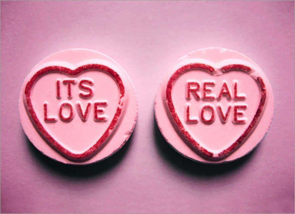 Cute Love Heart 桌布 壁紙 — 兩顆粉紅色心形糖果，帶有紅色閃粉邊緣，上面寫著「ITS LOVE」和「REAL LOVE」，並排放在柔軟的粉紅色表面上。