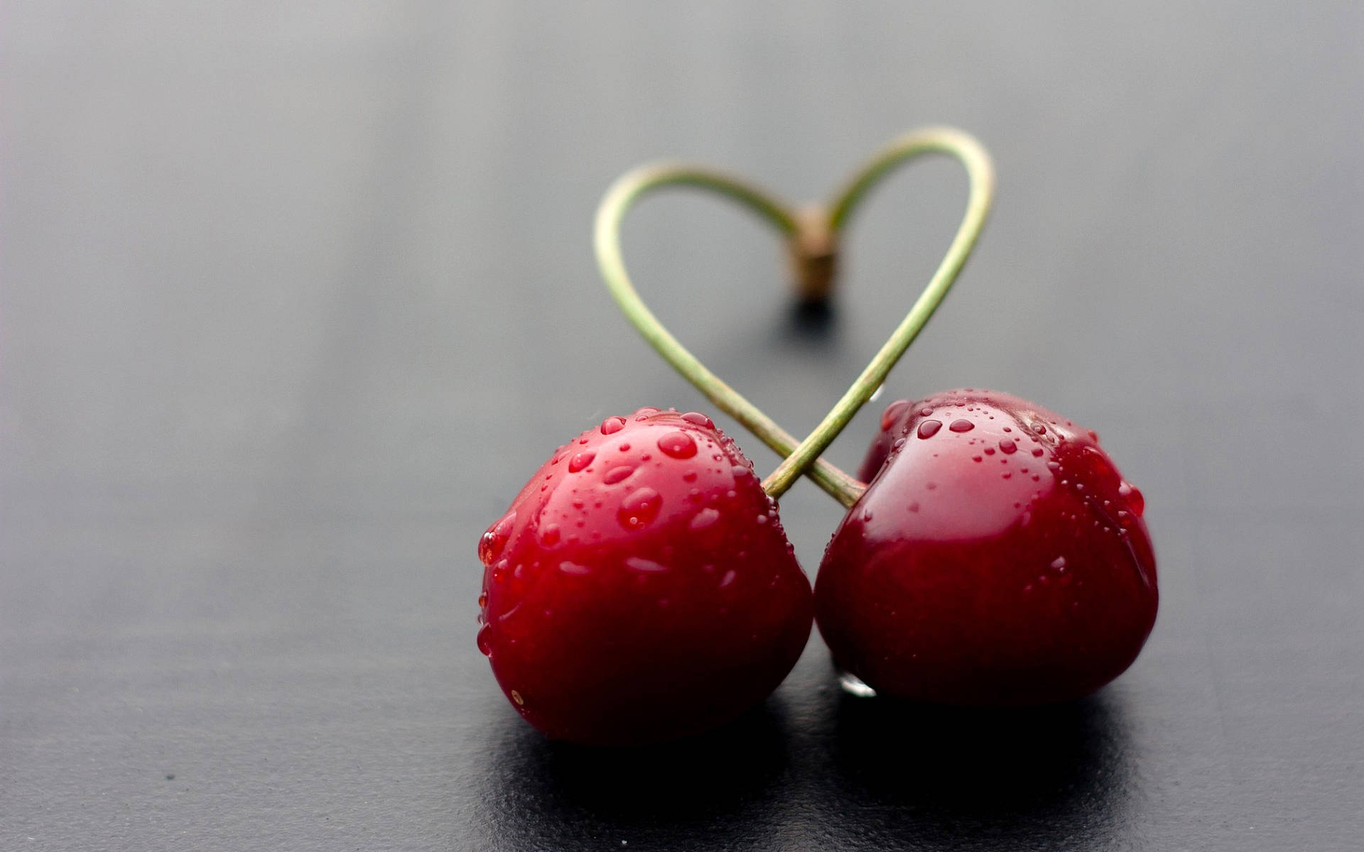 Cherry 桌布 壁紙 — 兩顆閃亮的紅櫻桃，莖部形成心形，置於深灰色表面上。