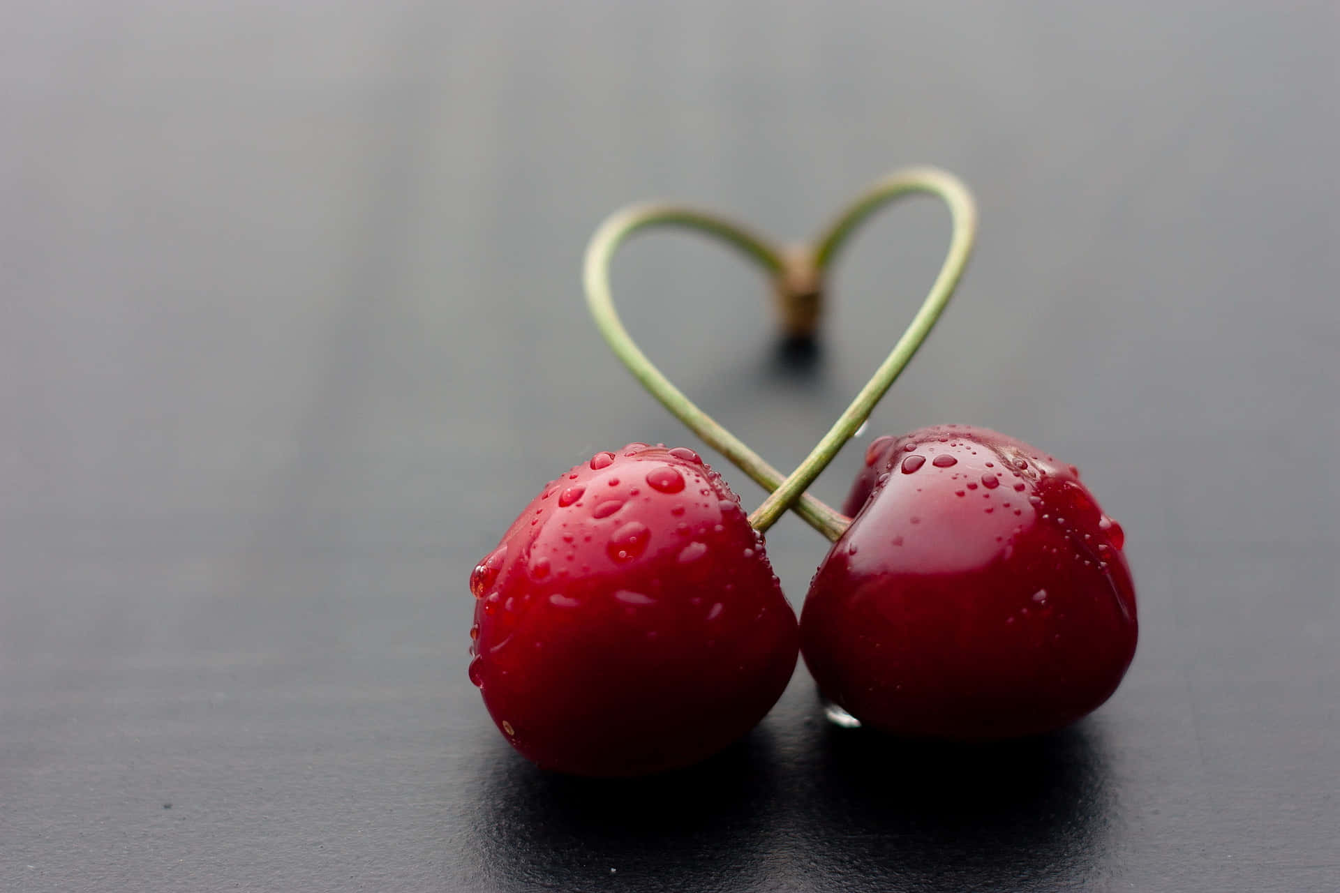 Kawaii Fruit 桌布 壁紙 — 兩顆閃亮的紅櫻桃，莖部形成心形，放在深灰色表面上。