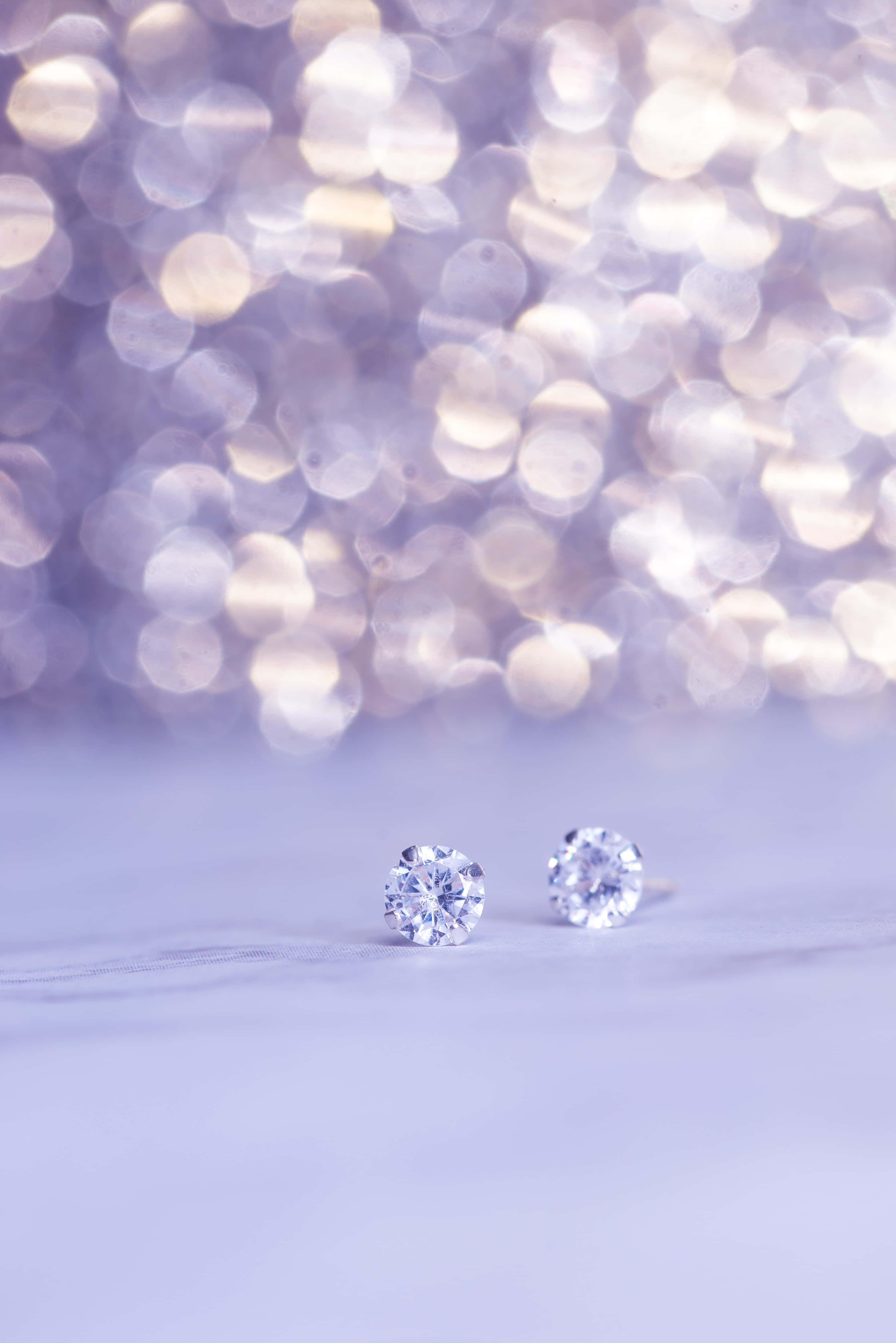 Earrings 桌布 壁紙 — 兩顆閃爍的鑽石耳釘放在柔軟的薰衣草色表面上，背景是閃爍燈光的夢幻散景。