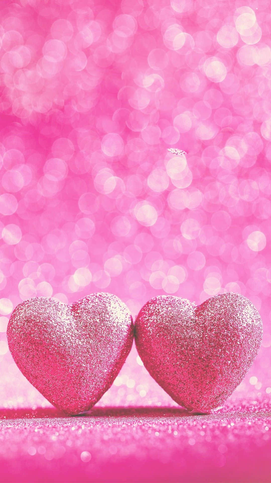 Glitter Pink Hearts 桌布 壁紙 — 兩顆閃閃發光的粉紅色心形並排放在閃爍的粉紅色表面上，背景為散景效果。