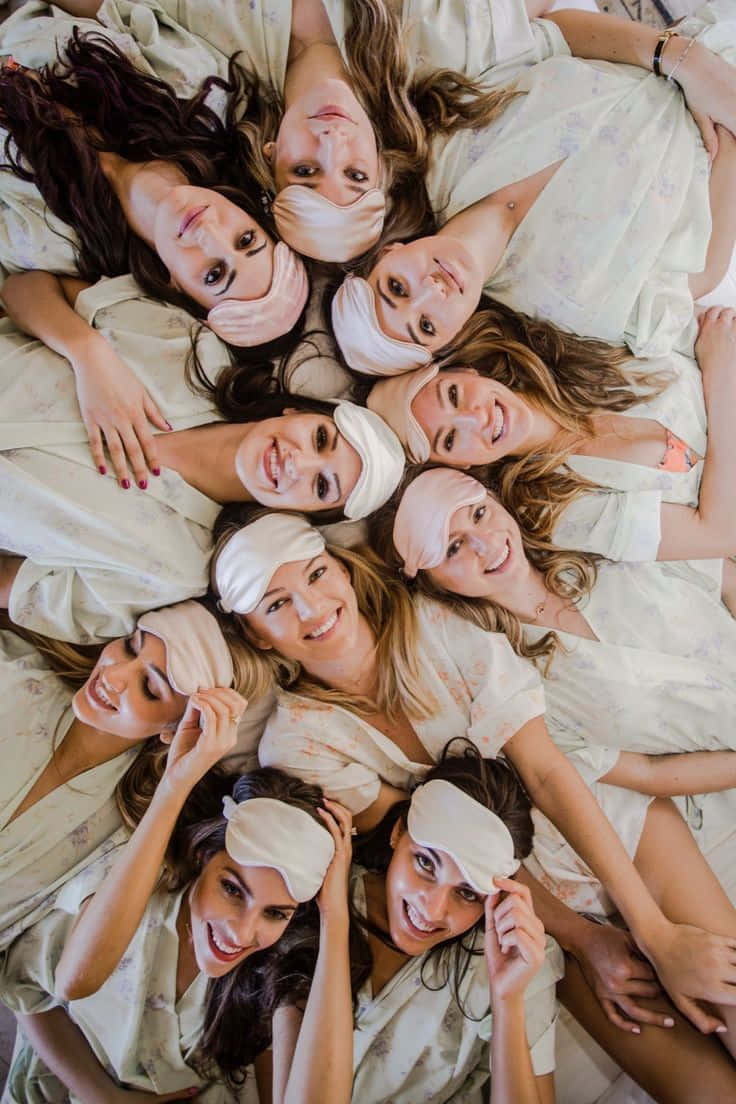 Bridesmaids 桌布 壁紙 — 八位微笑的女性穿著相配的淺色睡衣和眼罩，在床上圍成一圈躺著的俯視圖。