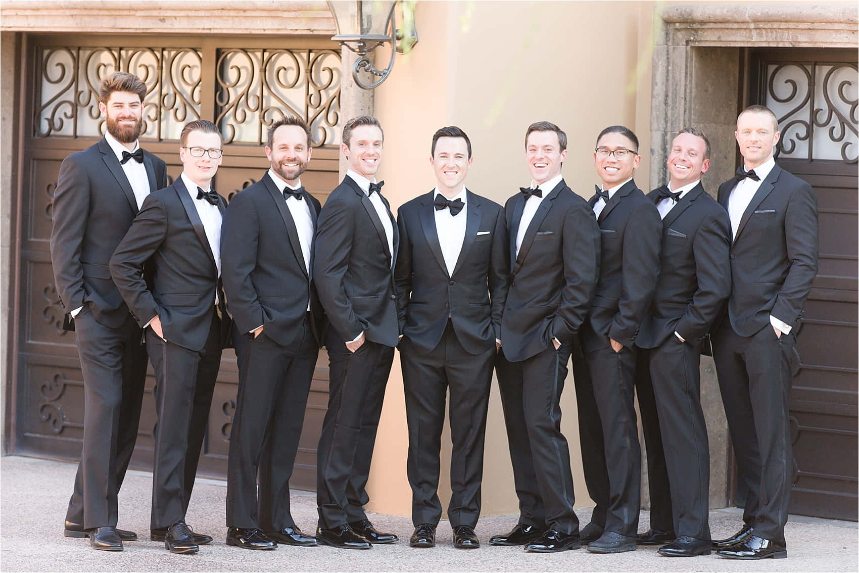 Groomsmen 桌布 壁紙 — 八個微笑的男人穿著黑色燕尾服和領結，在華麗的棕色門前自信地擺姿勢。