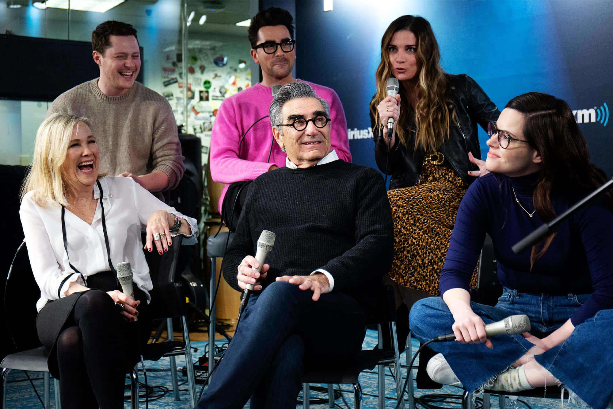 Eugene Levy 桌布 壁紙 — 六人在現代錄音室中，在麥克風和品牌背景前，於一場生動的播客錄製中大笑和聊天。