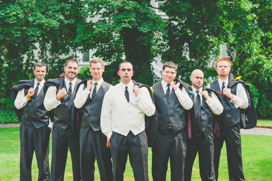 Groomsmen 桌布 壁紙 — 六位伴郎穿著黑色背心和白色襯衫，在戶外將外套搭在肩上，以茂密的綠樹為背景擺姿勢。