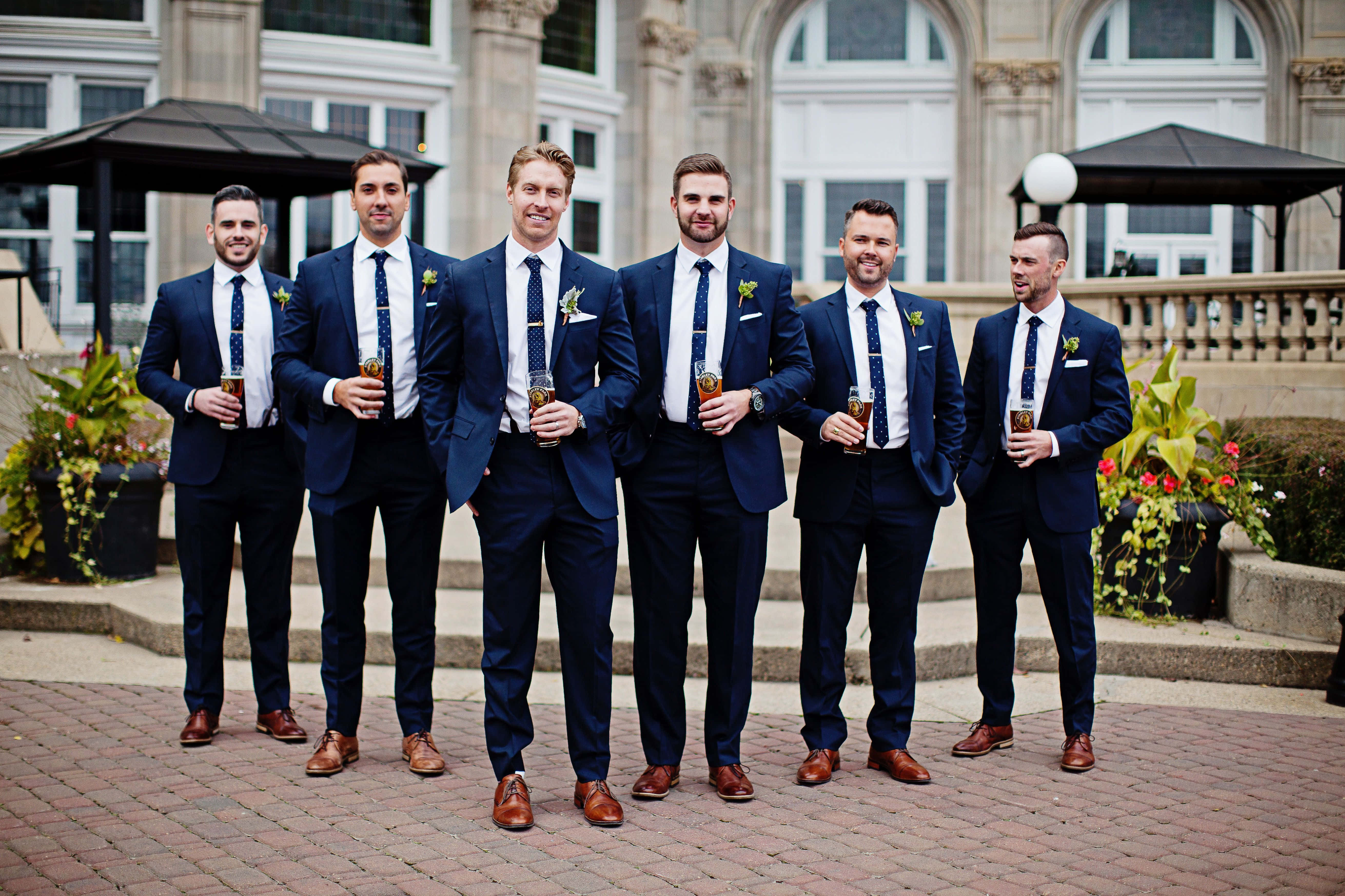 Groomsman 桌布 壁紙 — 六位伴郎身穿海軍藍西裝和圖案領帶，在宏偉建築前的磚階上微笑站立，手持飲品。