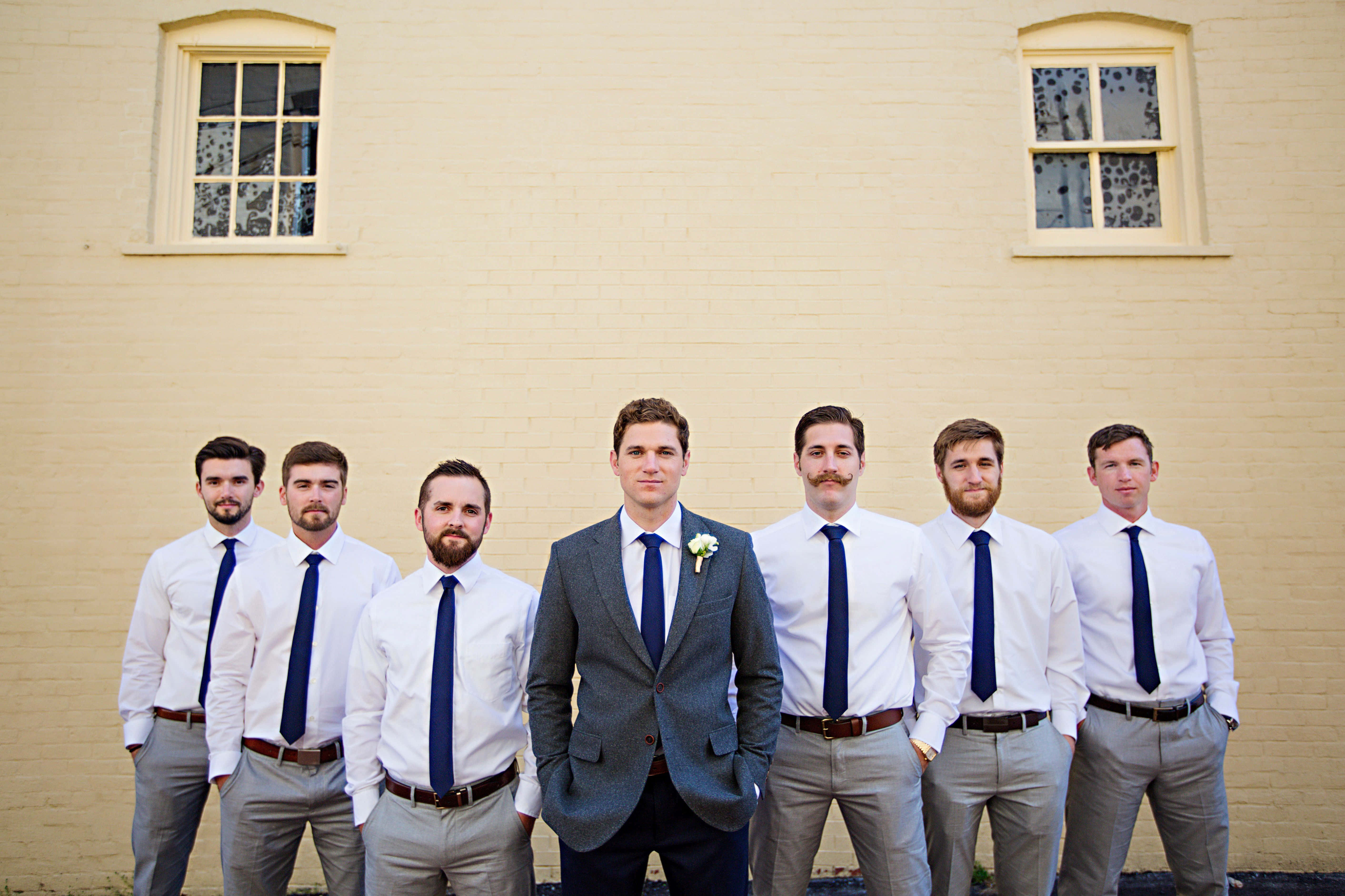 Groomsmen 桌布 壁紙 — 六位伴郎身穿白襯衫、海軍藍領帶和灰色長褲，站在一堵有兩扇窗戶的淡黃色磚牆前。