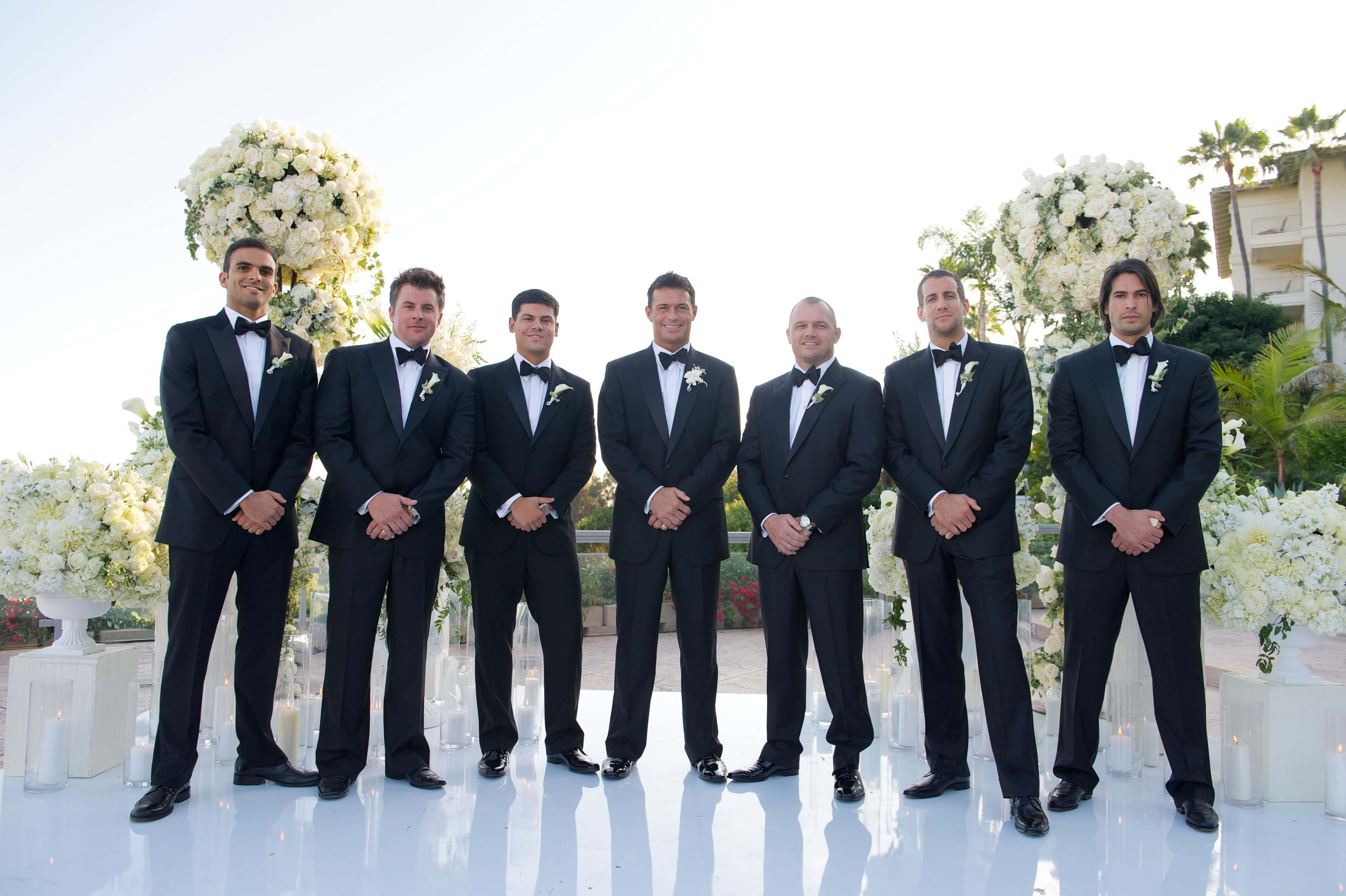 Groomsmen 桌布 壁紙 — 六位伴郎身穿黑色燕尾服，佩戴白色襟花，在明亮的日光下站成一排，位於白色反光平台上。