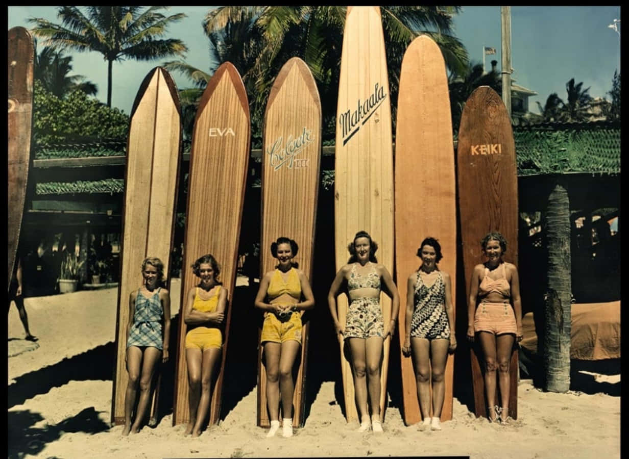 Vintage Surf 桌布 壁紙 — 六位復古女性穿著復古泳衣，在陽光明媚、棕櫚樹成蔭的海灘上自信地站在高大的木製衝浪板旁。