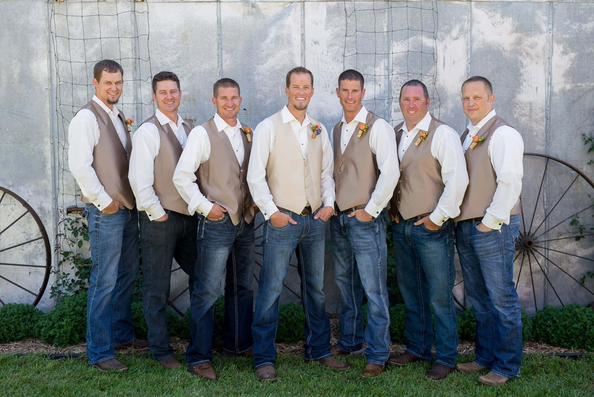 Groomsmen 桌布 壁紙 — 六位微笑的伴郎穿著相配的米色背心和藍色牛仔褲，在帶有馬車輪的質樸金屬柵欄前自信地擺姿勢。