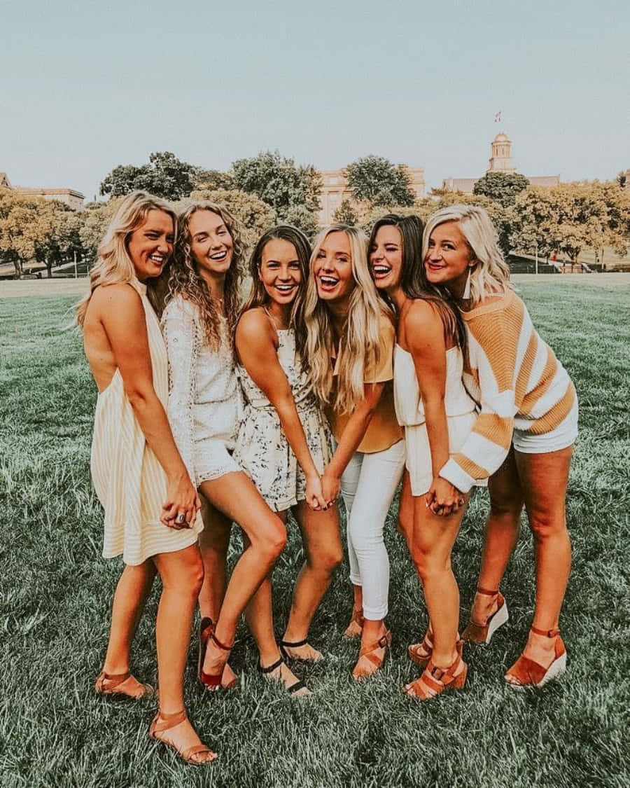 girls 桌布 壁紙 — 六位微笑的女性穿著夏季服裝，在草地上一起大笑，草地有樹木和遠處的建築物，天空晴朗。