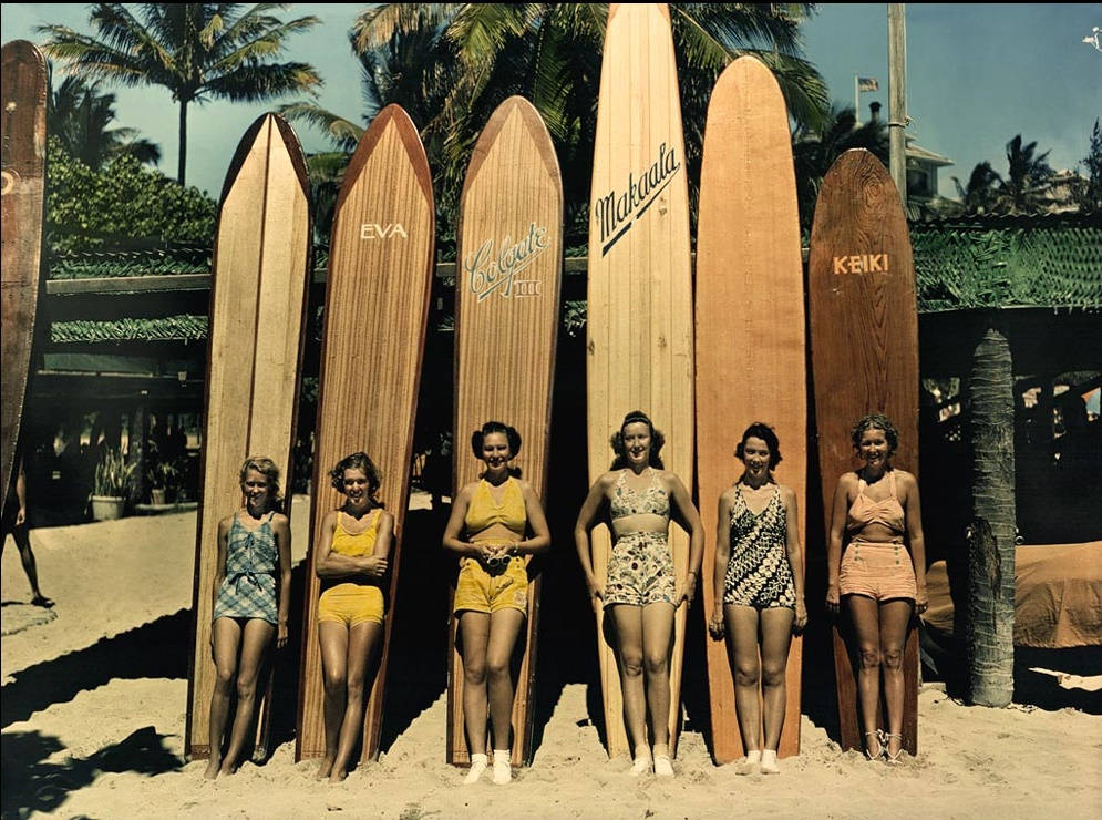 Surfboard 桌布 壁紙 — 六位微笑的女性穿著復古泳衣，在陽光明媚的海灘上，棕櫚樹旁，與高大的木製衝浪板合影。