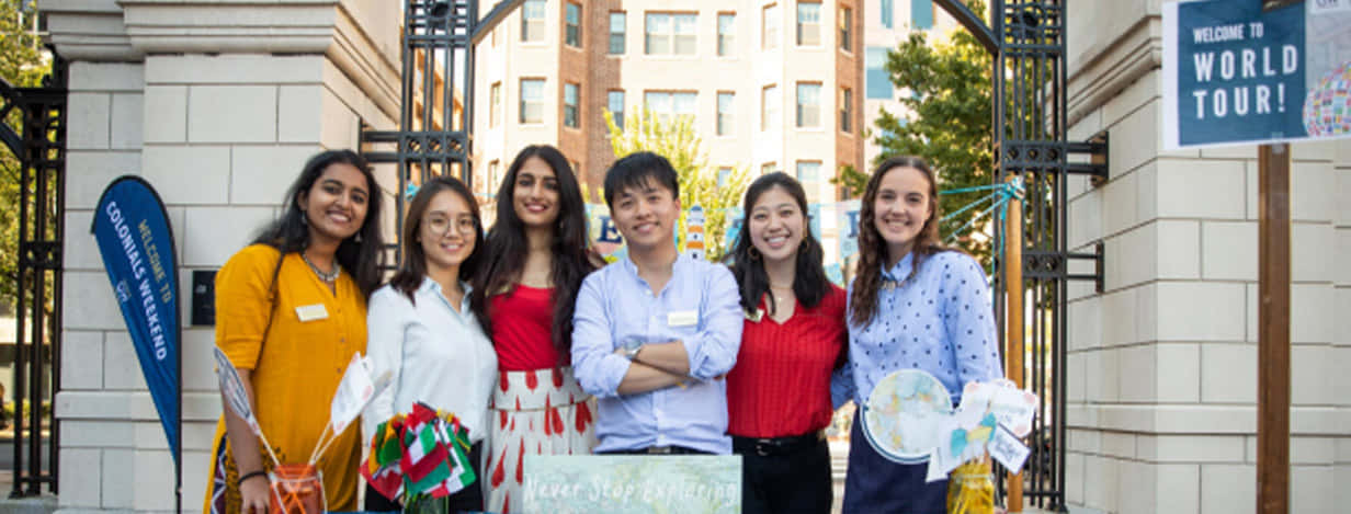 學校 桌布 壁紙 — 六位微笑的年輕成年人在大學門口愉快地擺姿勢，手持旅行主題道具，在明亮的日光下。