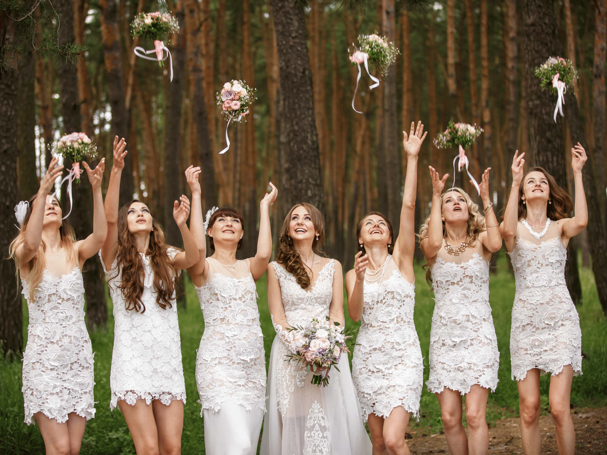 Bridesmaids 桌布 壁紙 — 六位快樂的伴娘和一位身穿白色蕾絲連衣裙的新娘在陽光明媚的松樹林中向上拋擲花束。