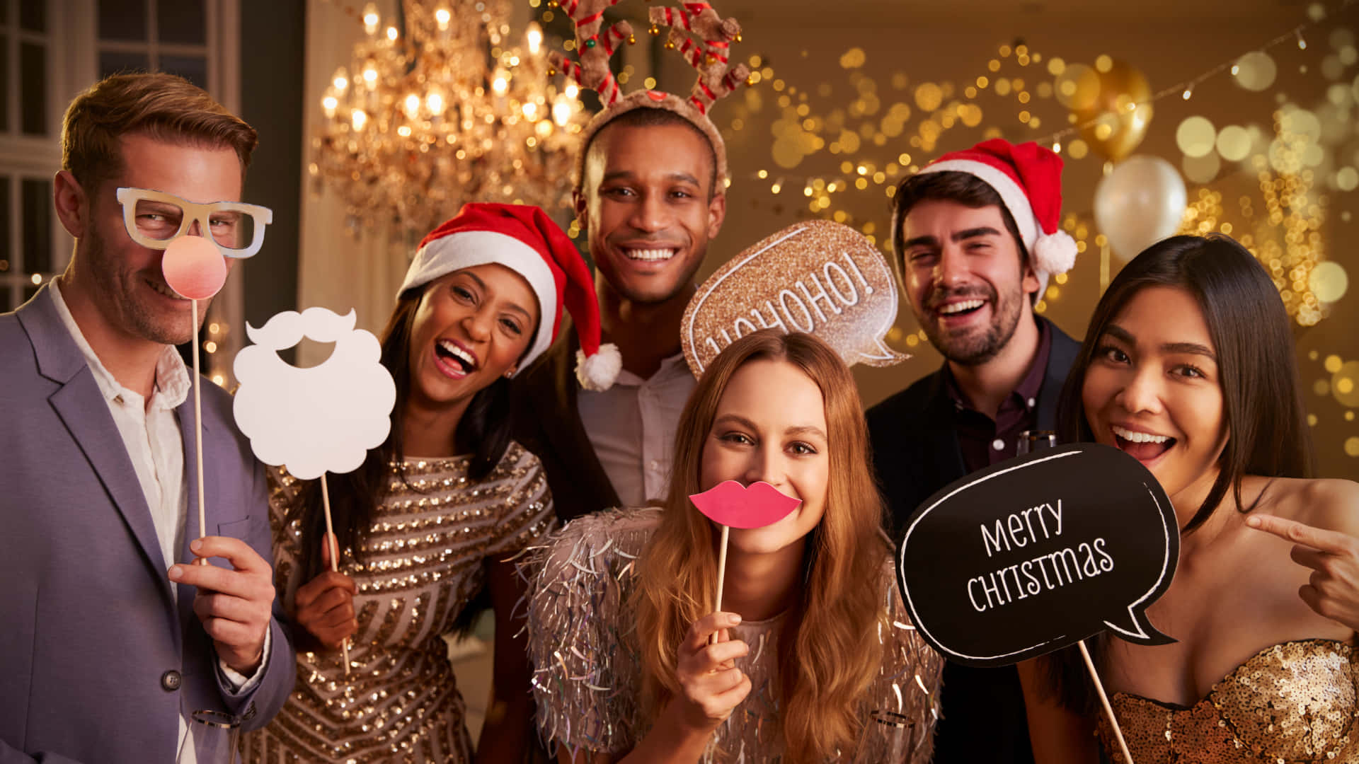 Christmas Virtual 桌布 壁紙 — 六位快樂的朋友穿著節日服裝，在溫暖的金色燈光和閃爍的水晶吊燈下，拿著有趣的道具擺姿勢。