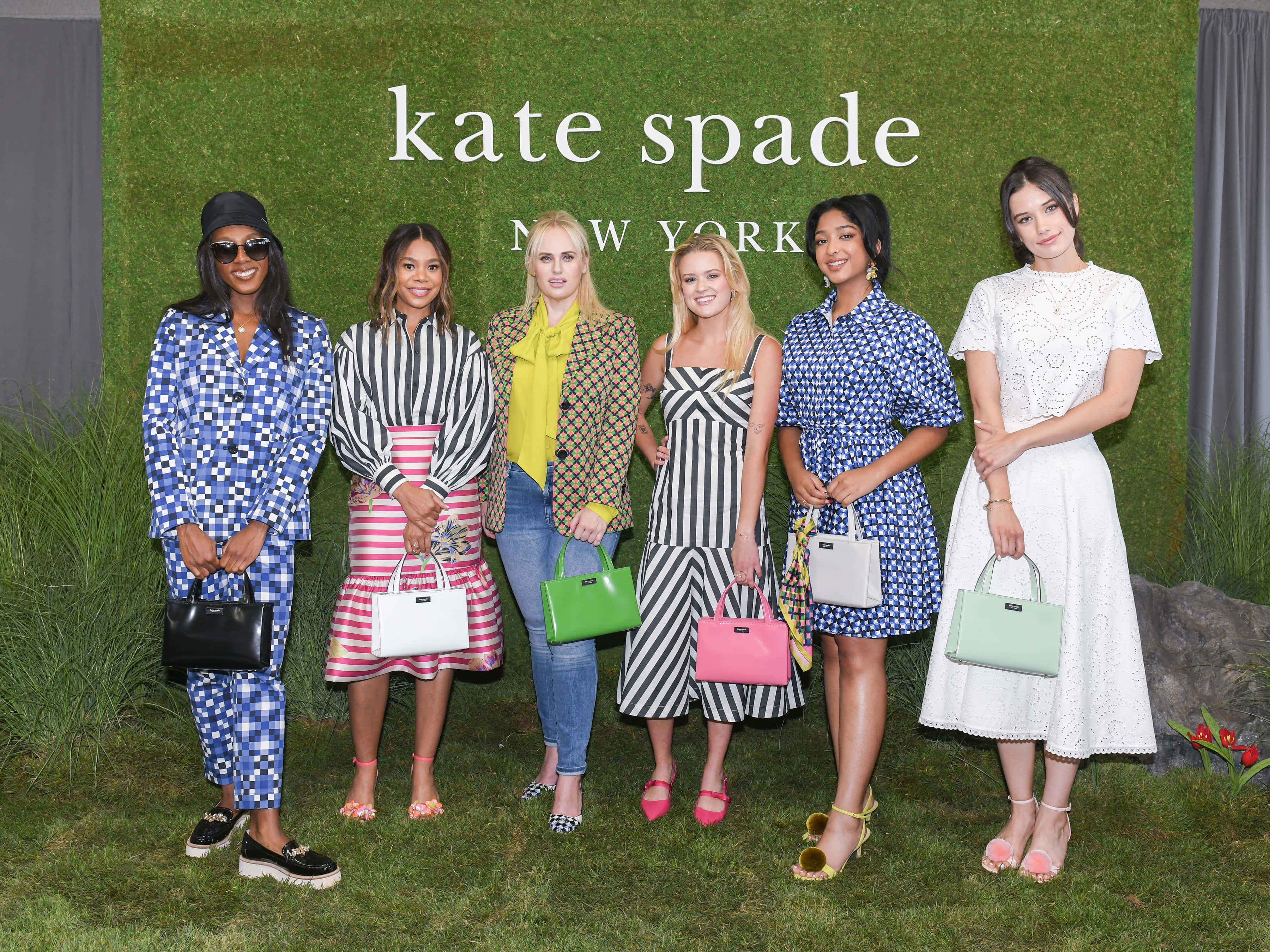 kate spade 桌布 壁紙 — 六位時尚女性穿著色彩繽紛的圖案服裝和手提包，在帶有 kate spade new york 品牌的綠色苔蘚牆前擺姿勢。