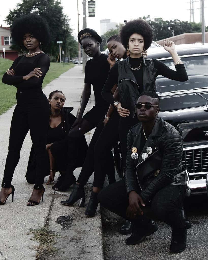 Black Panther Party 桌布 壁紙 — 六位時尚的黑人個體穿著黑色服裝，在多雲的天空下，自信地在復古車旁的人行道上擺姿勢。