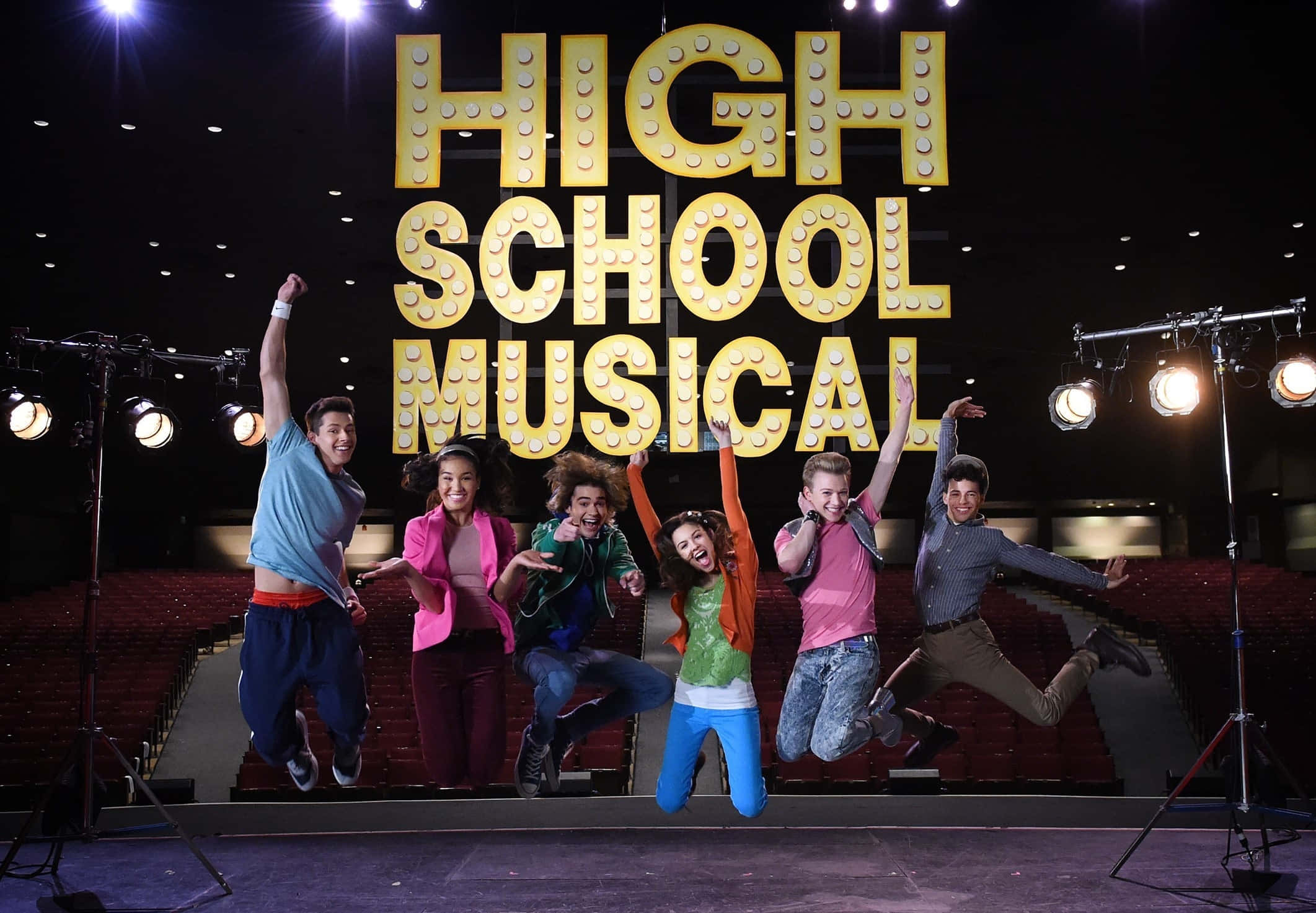 High School Musical 桌布 壁紙 — 六位歡樂的演員在發光的「HIGH SCHOOL MUSICAL」招牌下，在劇院舞台上充滿活力地跳躍。