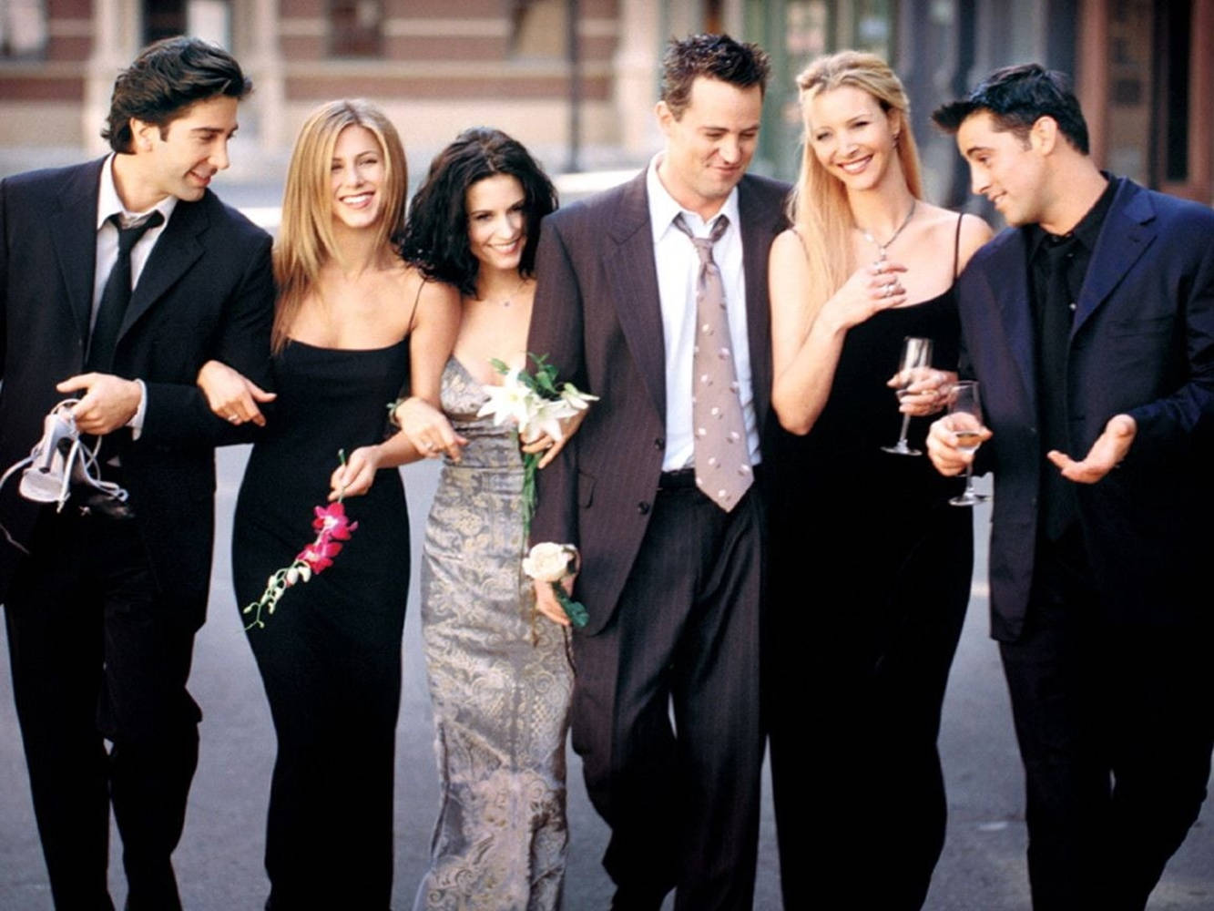 Friends Aesthetic 桌布 壁紙 — 六位穿著正式服裝的朋友笑著一起走在城市街道上，手持飲料和鮮花。