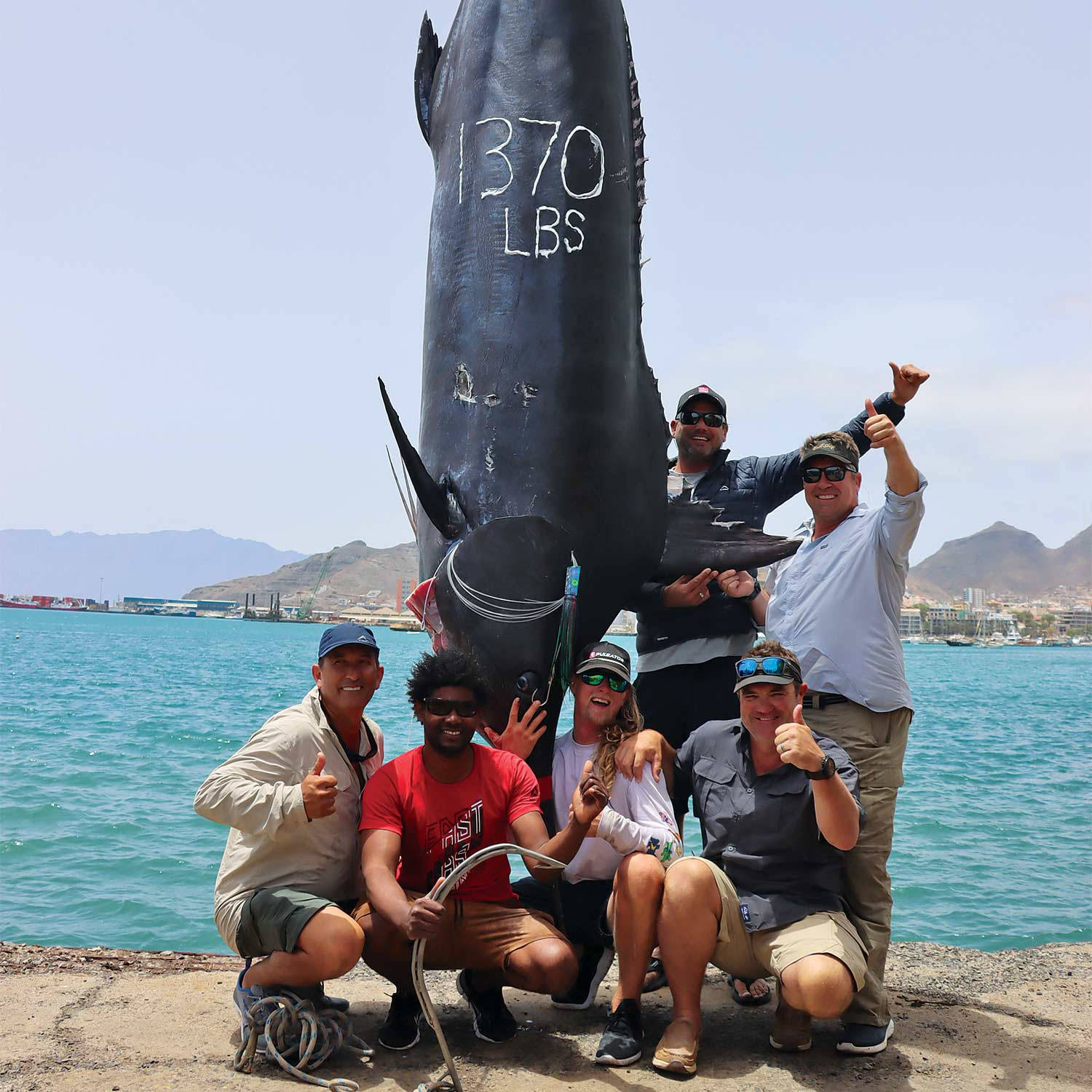 六位開心的釣魚人士在陽光明媚的碼頭上慶祝捕獲一條標示為1370磅的巨大鮪魚。 — 釣魚 GIF 壁紙