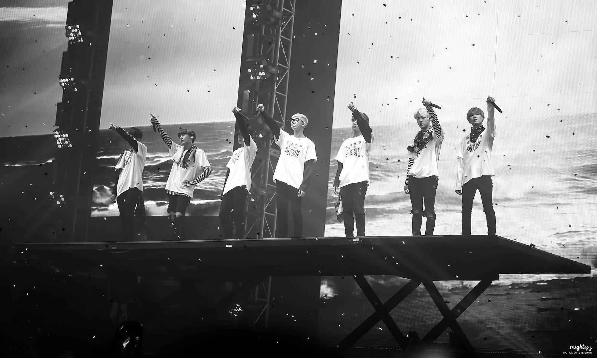 BTS 桌布 壁紙 — 六位表演者在升高的舞台上，在單色背景下，齊聲舉起手臂，背景是洶湧的海浪和飄落的五彩紙屑。