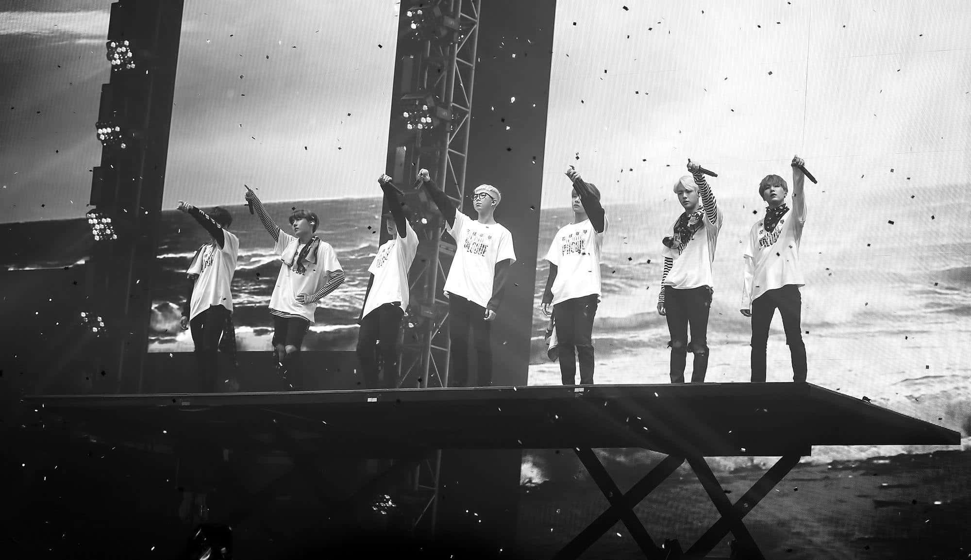 BTS 美學 桌布 壁紙 — 六位表演者在黑白的高架舞台上，在明亮的舞台燈光下，於飄落的五彩紙屑中舉起雙臂。