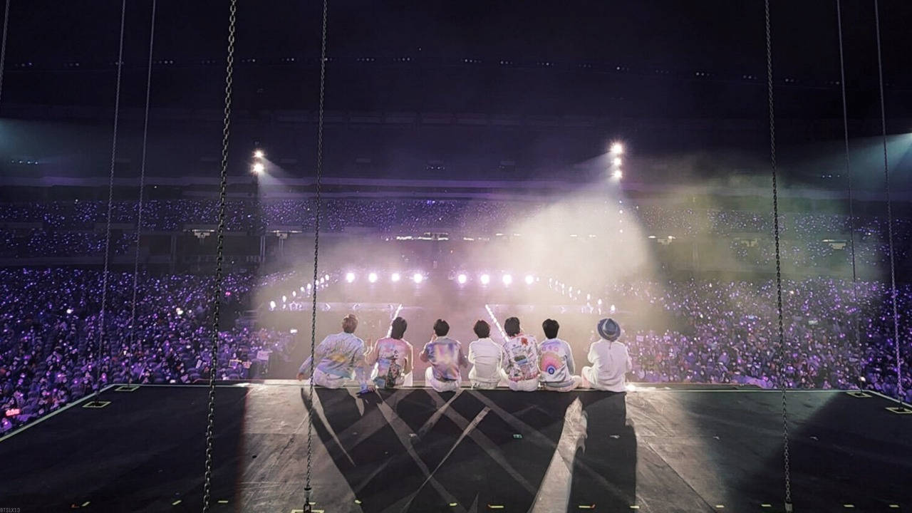 六位表演者坐在懸掛舞台上，面向夜間手持紫色燈光的大量人群。 — BTS ARMY GIF 壁紙