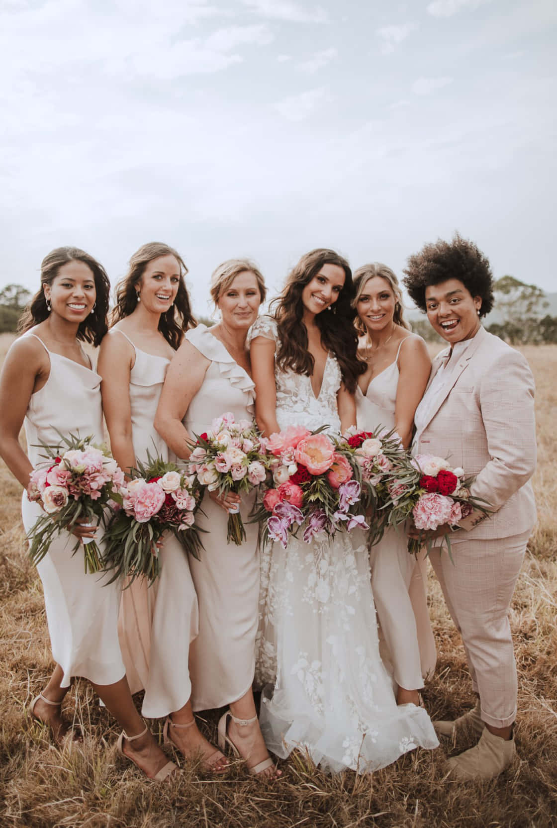 Bridal 桌布 壁紙 — 六位身穿柔和米色和白色服裝的女士組成的歡樂婚禮派對，一起站在陽光明媚的田野中，手持充滿活力的粉紅色和紅色花束。