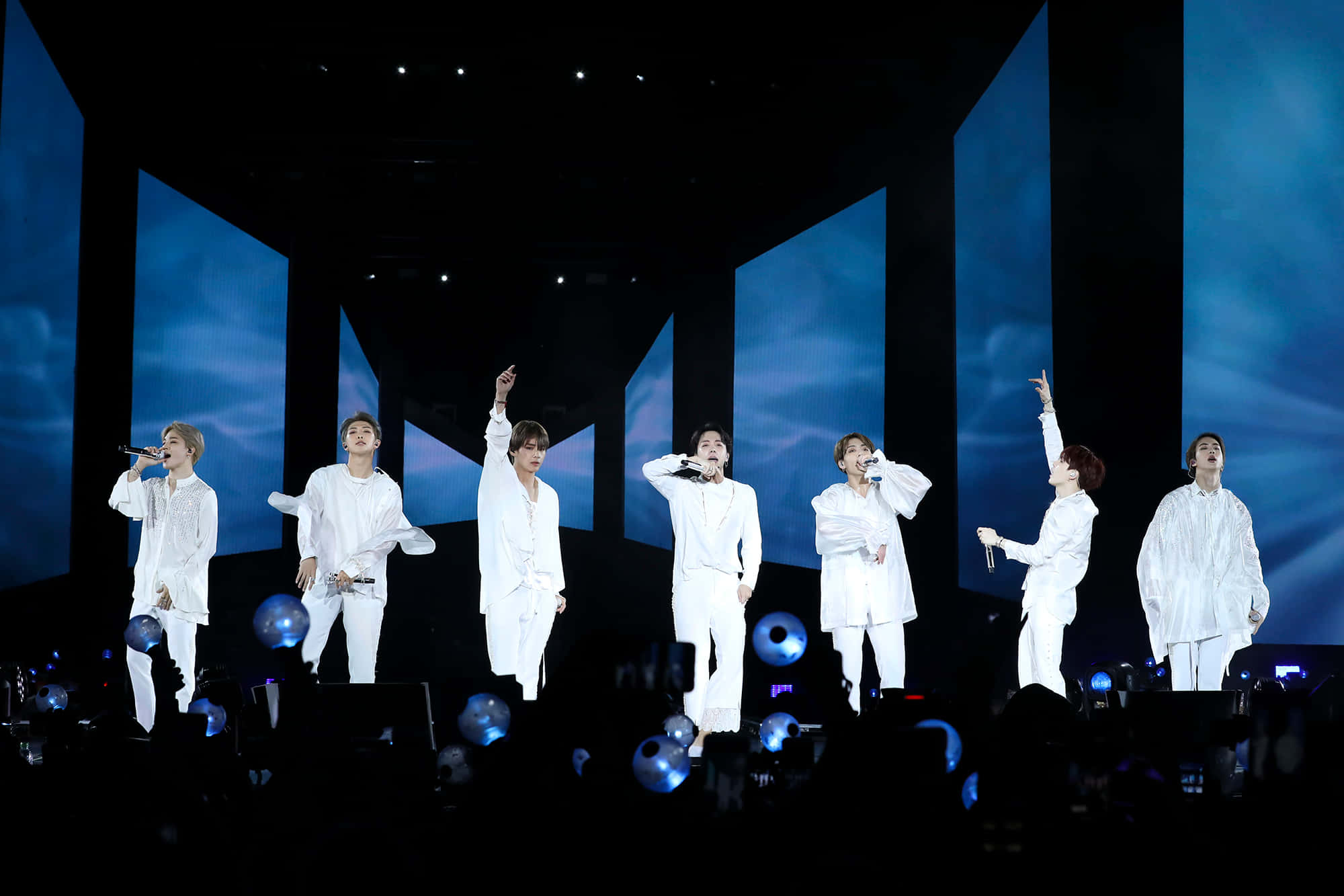 Bts Stage 桌布 壁紙 — 六位身穿白色服裝的男表演者在一個帶有藍色垂直螢幕和發光球體的黑暗舞台上唱歌。