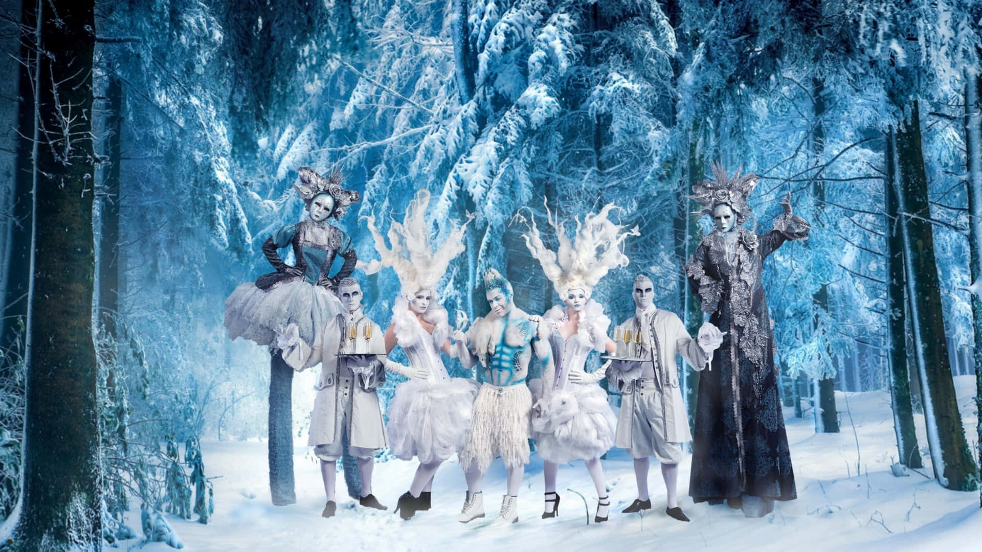 winter wonderland 桌布 壁紙 — 六位身穿精緻服裝、化著戲劇性妝容、戴著羽毛頭飾的表演者在藍色調的天空下，於雪地森林中擺姿勢。