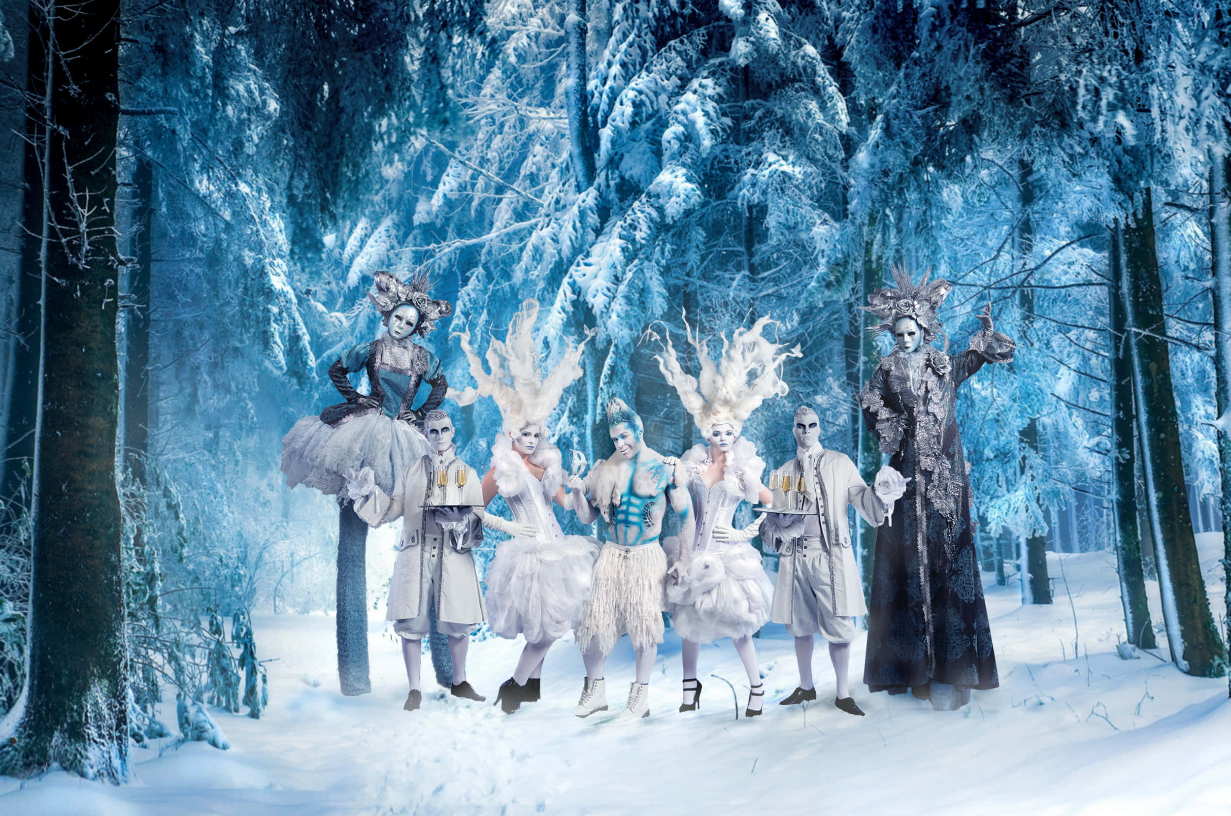 Christmas Winter Wonderland 桌布 壁紙 — 六位身穿精緻服裝、化著戲劇性白色妝容、戴著羽毛頭飾的表演者，在沐浴在冰冷藍光中的雪地森林裡擺姿勢。