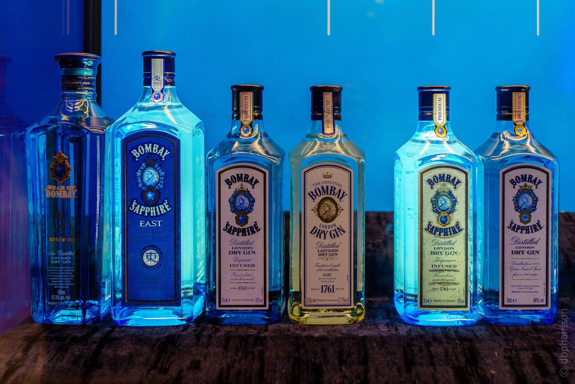  壁紙 — 六個 Bombay Sapphire 琴酒瓶排列在木質表面上，在藍色燈光下，標籤和液體顏色各異。