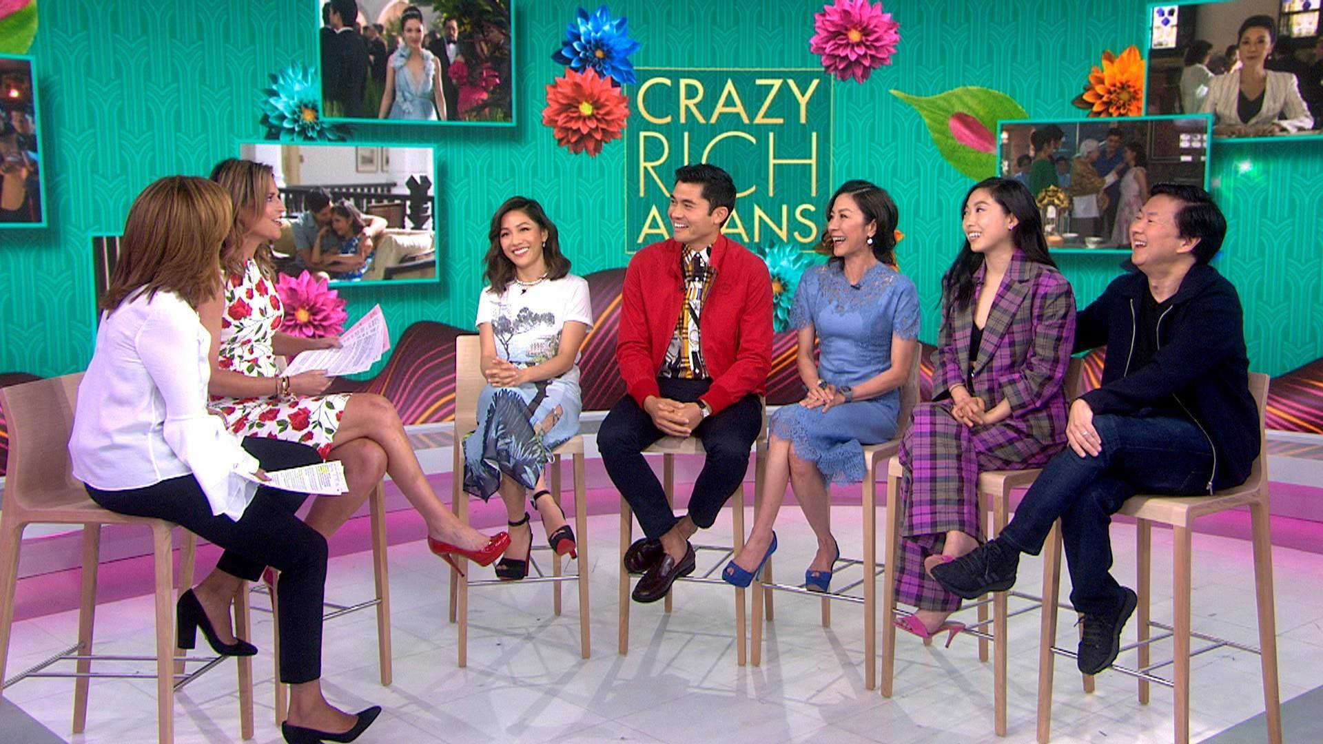 Rich 桌布 壁紙 — 六個人在色彩繽紛的花卉裝飾和「Crazy Rich Asians」標誌的充滿活力的電視攝影棚場景中，坐在凳子上大笑和聊天。