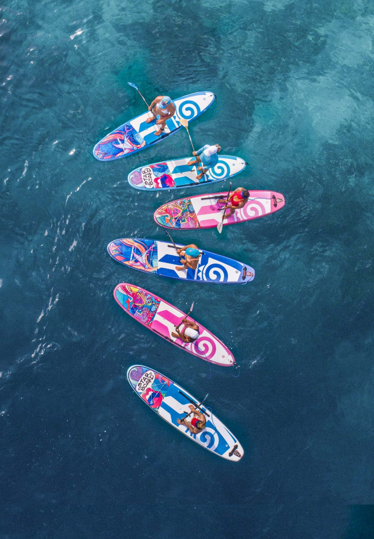 Paddleboarding 桌布 壁紙 — 六個人踩著色彩繽紛的槳板在深藍色海洋上滑行的鳥瞰圖。