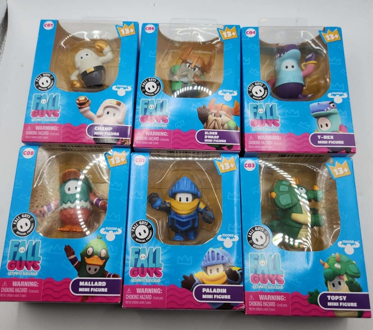 toys 桌布 壁紙 — 六個包裝好的 Fall Guys 迷你人偶展示在色彩繽紛的零售盒中，放在純白色表面上。