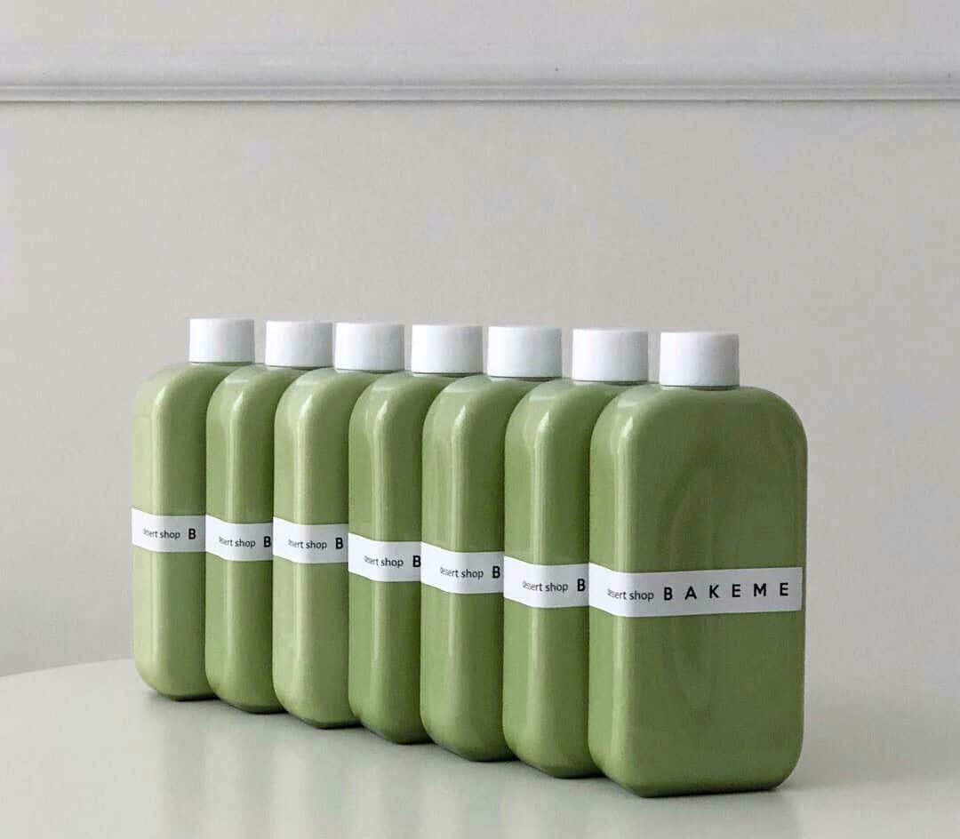 Aesthetic Sage Green 桌布 壁紙 — 六個啞光橄欖綠色瓶子，帶白色蓋子和標籤，整齊地排成一排，放在白色表面上，背景是素牆。