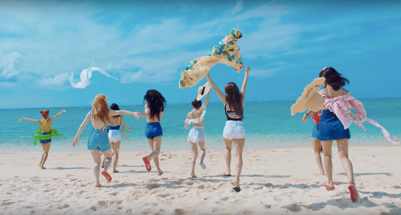 twice 桌布 壁紙 — 六個女人在陽光明媚的海灘上快樂地奔跑，將彩色毛巾拋向空中，背景是明亮的藍天。
