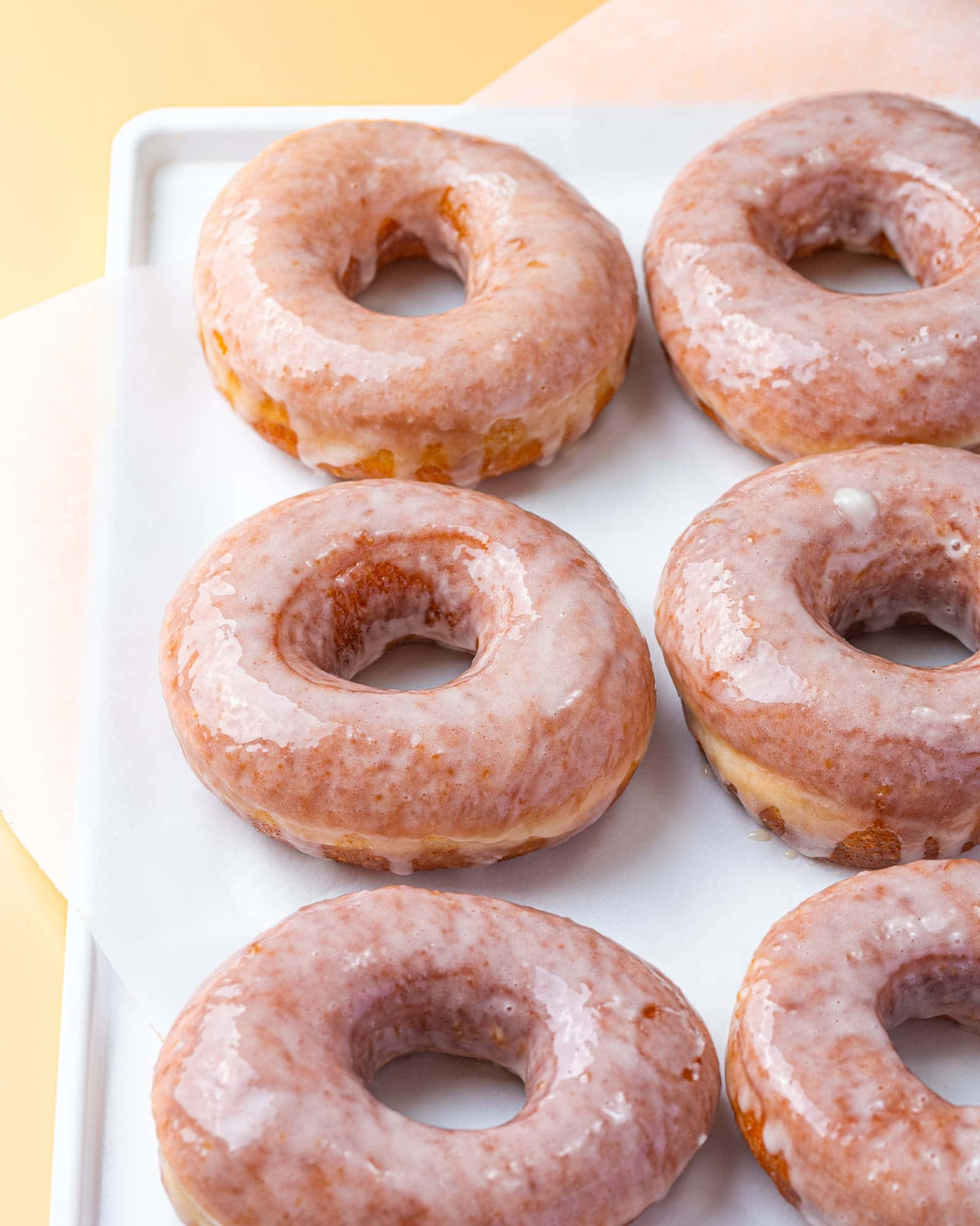 Glazed Donut 桌布 壁紙 — 六個帶有閃亮粉紅色糖霜的釉面甜甜圈整齊地放在白色托盤上，背景是柔和的黃色。