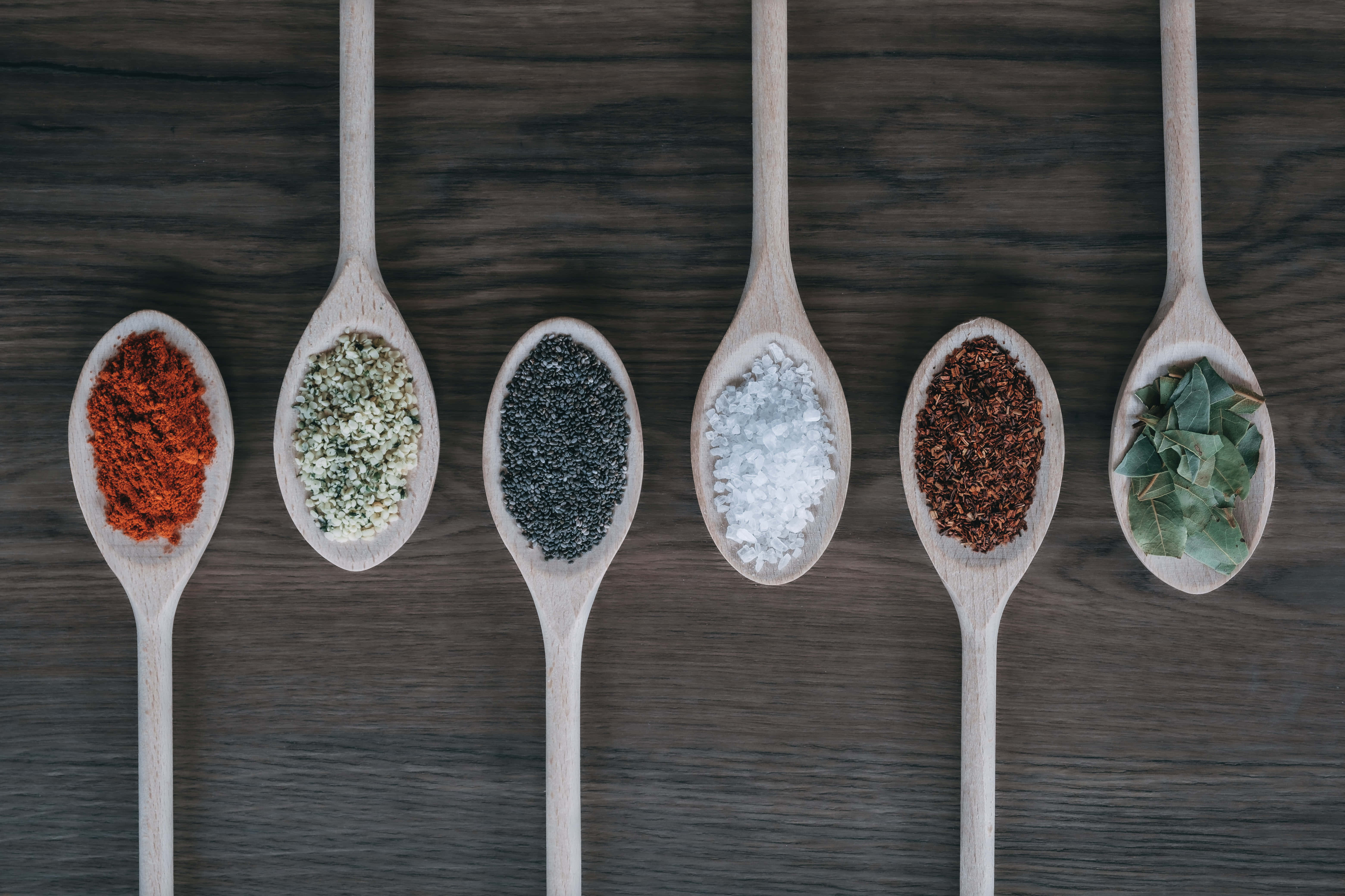Spices 桌布 壁紙 — 六個木勺盛載著色彩鮮豔的香料和香草，整齊地排列在深色木質表面上。