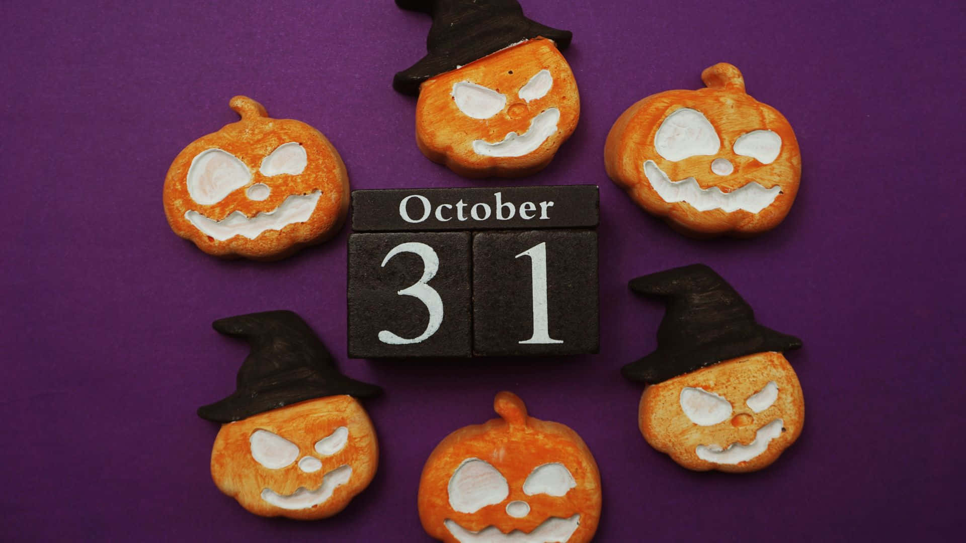 October 31st 桌布 壁紙 — 六個橙色南瓜形狀的餅乾，帶有白色糖霜臉和黑色巫師帽，圍繞著一個顯示10月31日的黑色木塊，背景為深紫色。