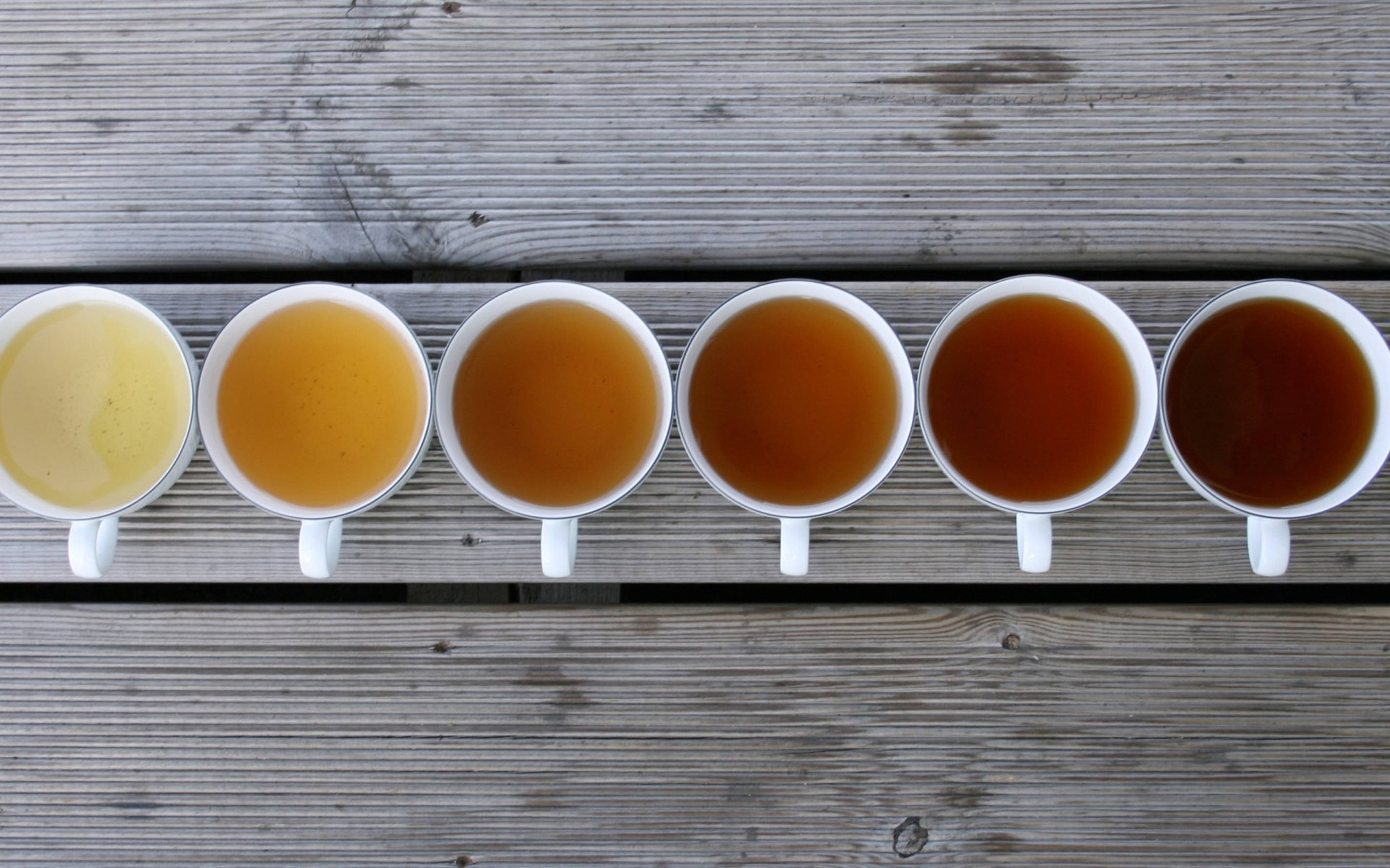 tea 桌布 壁紙 — 六個白色馬克杯裝滿了從淡黃色到深棕色的茶，排列在風化的木板上。